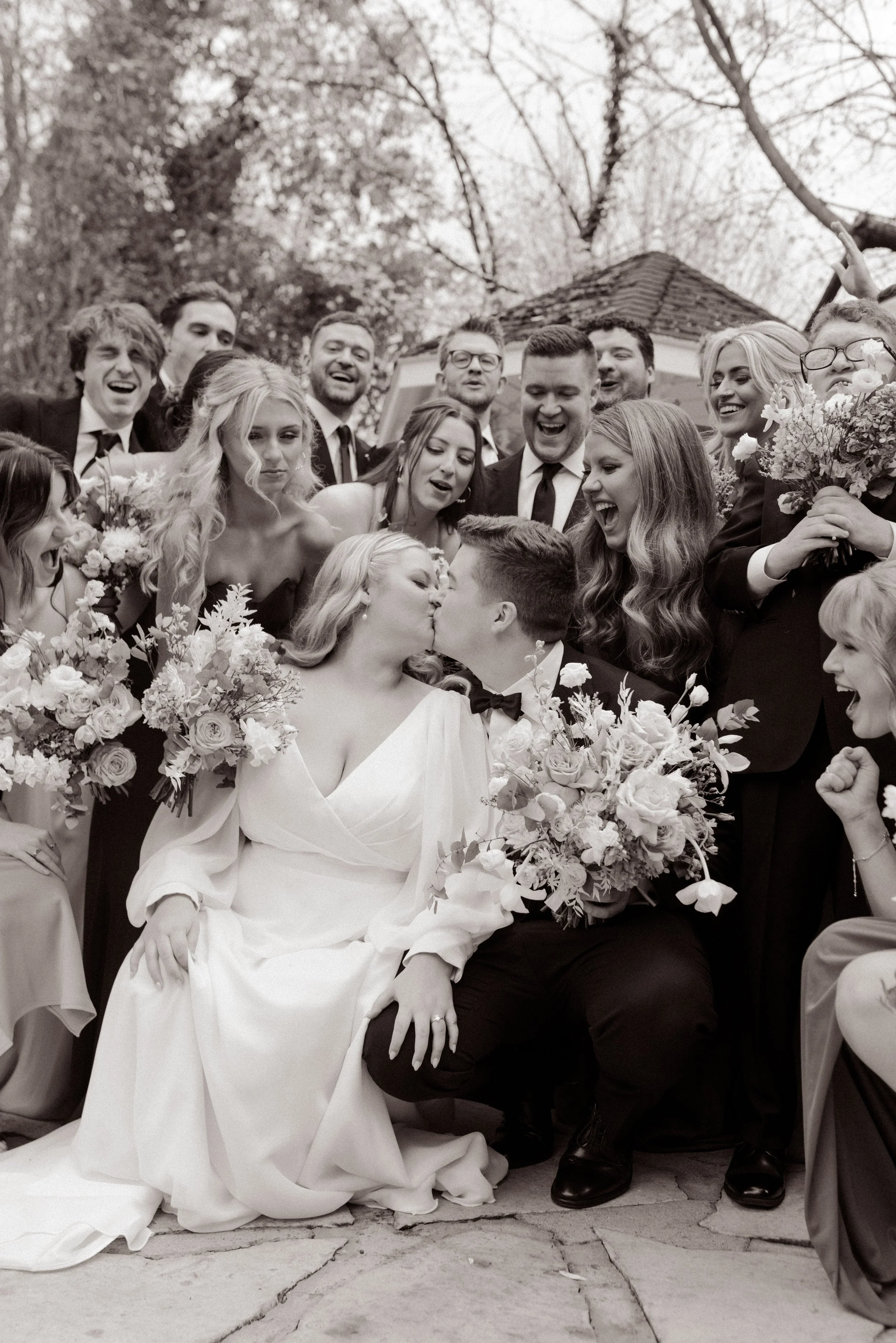 Black-and-white wedding party photo of the newlyweds kissing while their friends laugh and celebrate around them.