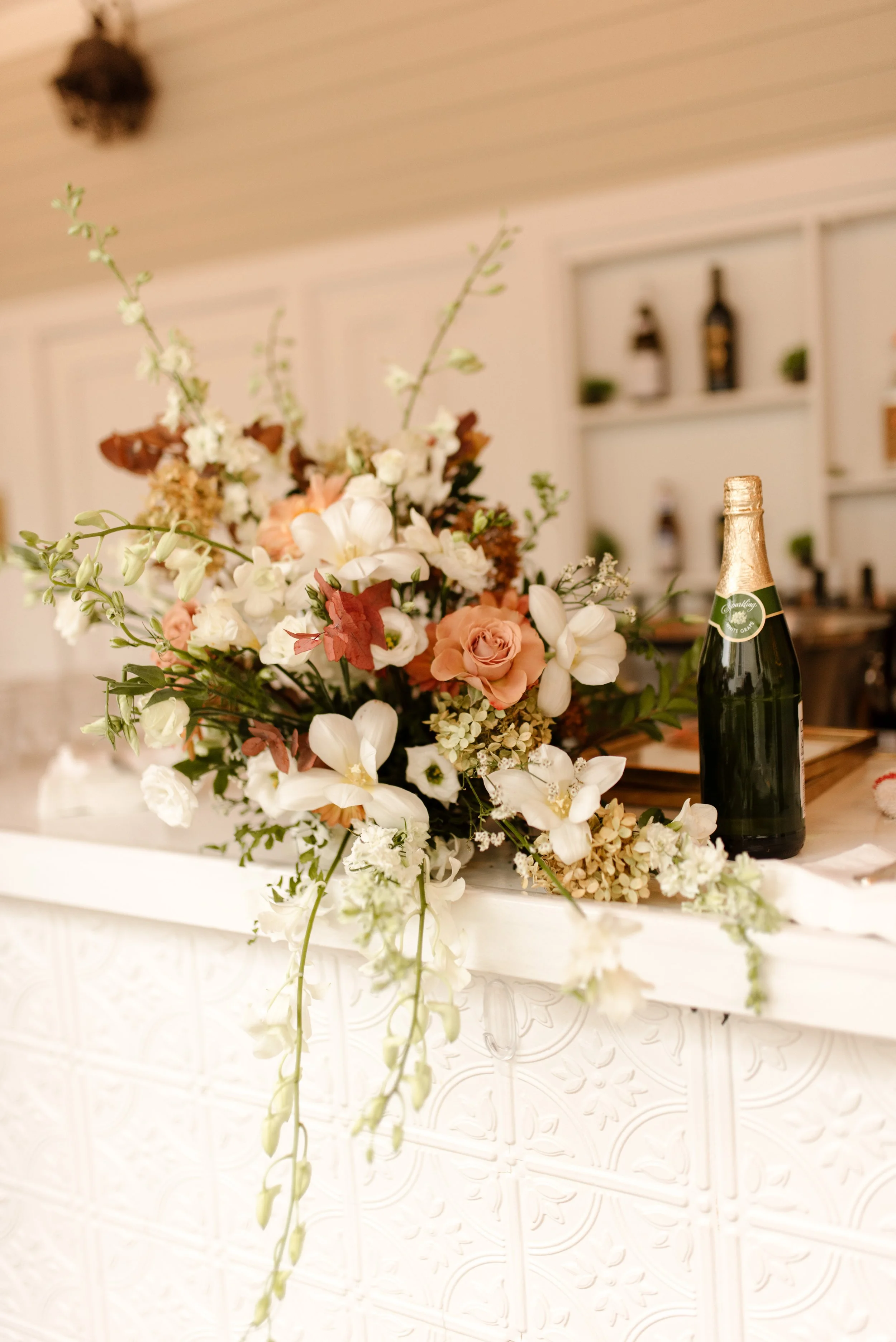 Peach, rust, and white floral arrangement beside a champagne bottle at the bar for a romantic fall wedding reception.