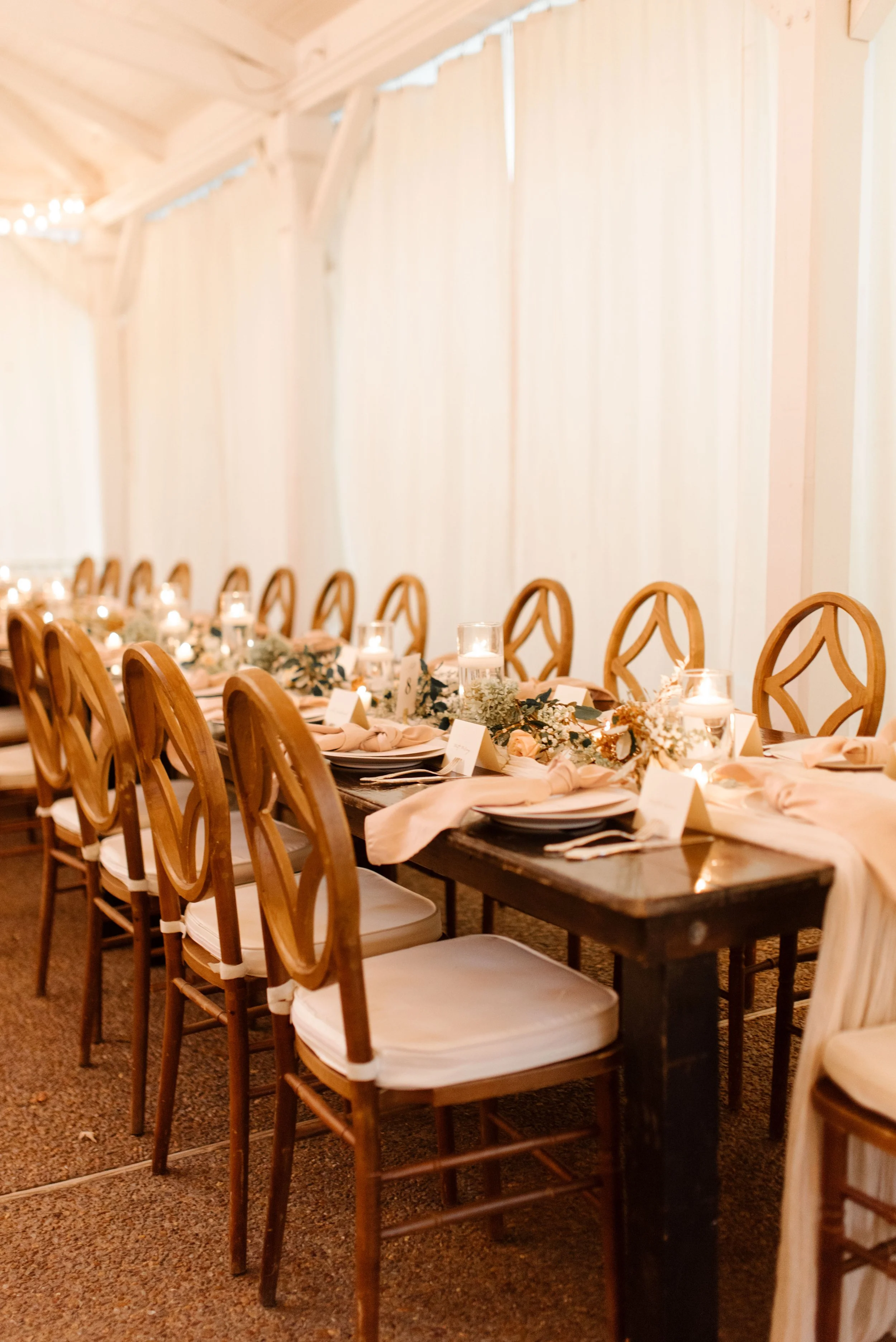 Long reception tables set with candles, greenery, neutral linens, and warm wood chairs beneath a white tent.