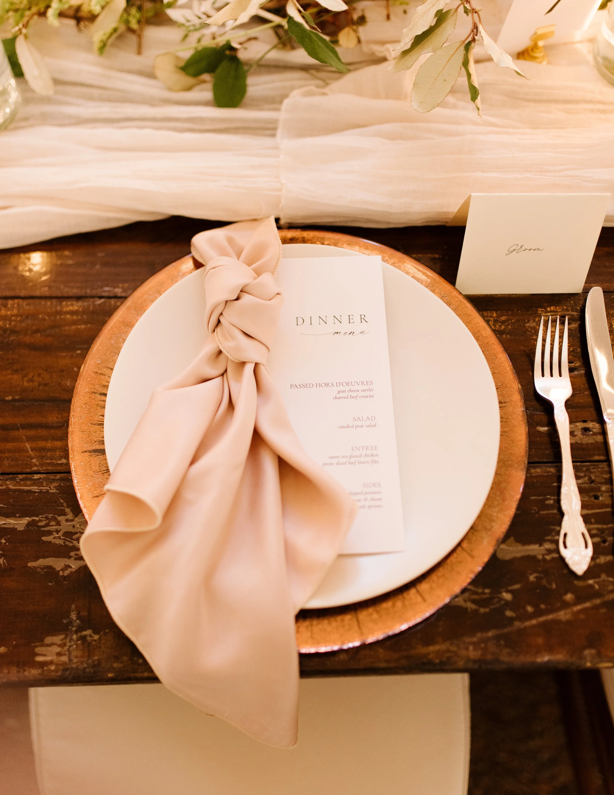 Fall wedding place setting with a blush napkin, dinner menu, gold charger, greenery, and personalized place card.