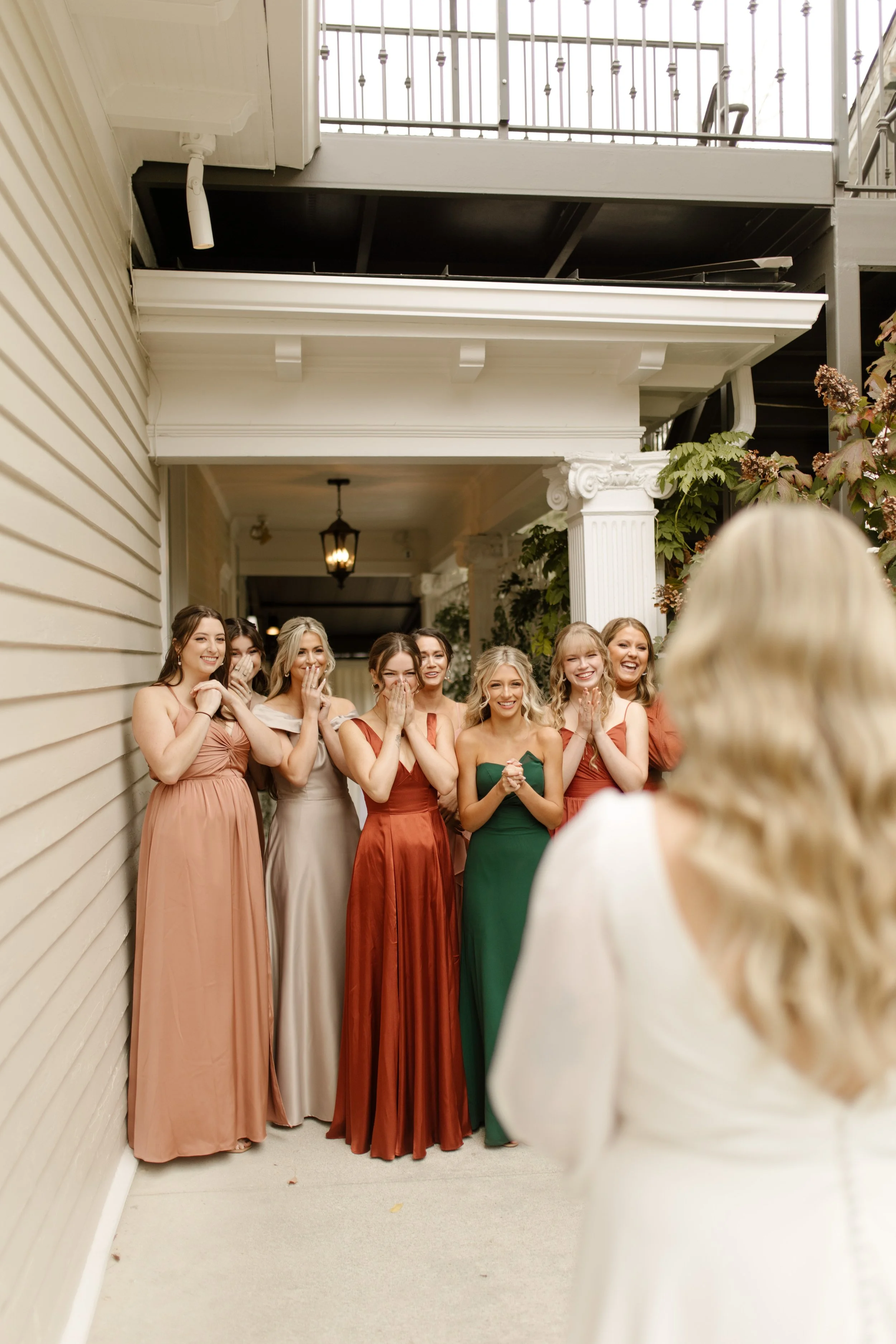 Bridesmaids in rust, green, blush, and neutral dresses reacting as they see the bride in her wedding dress.