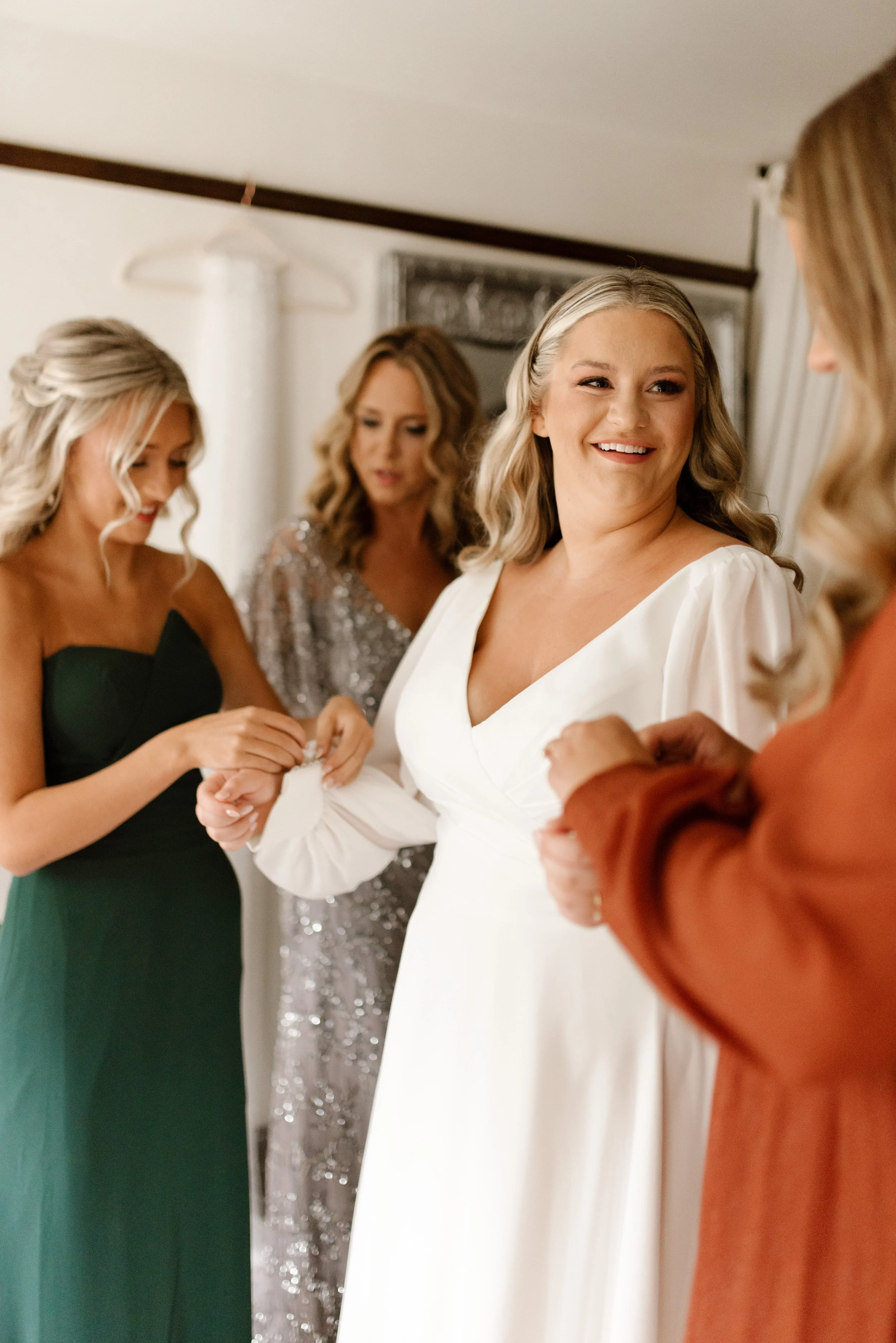 Bride smiling with her bridesmaids while getting ready in her long-sleeved white wedding dress.