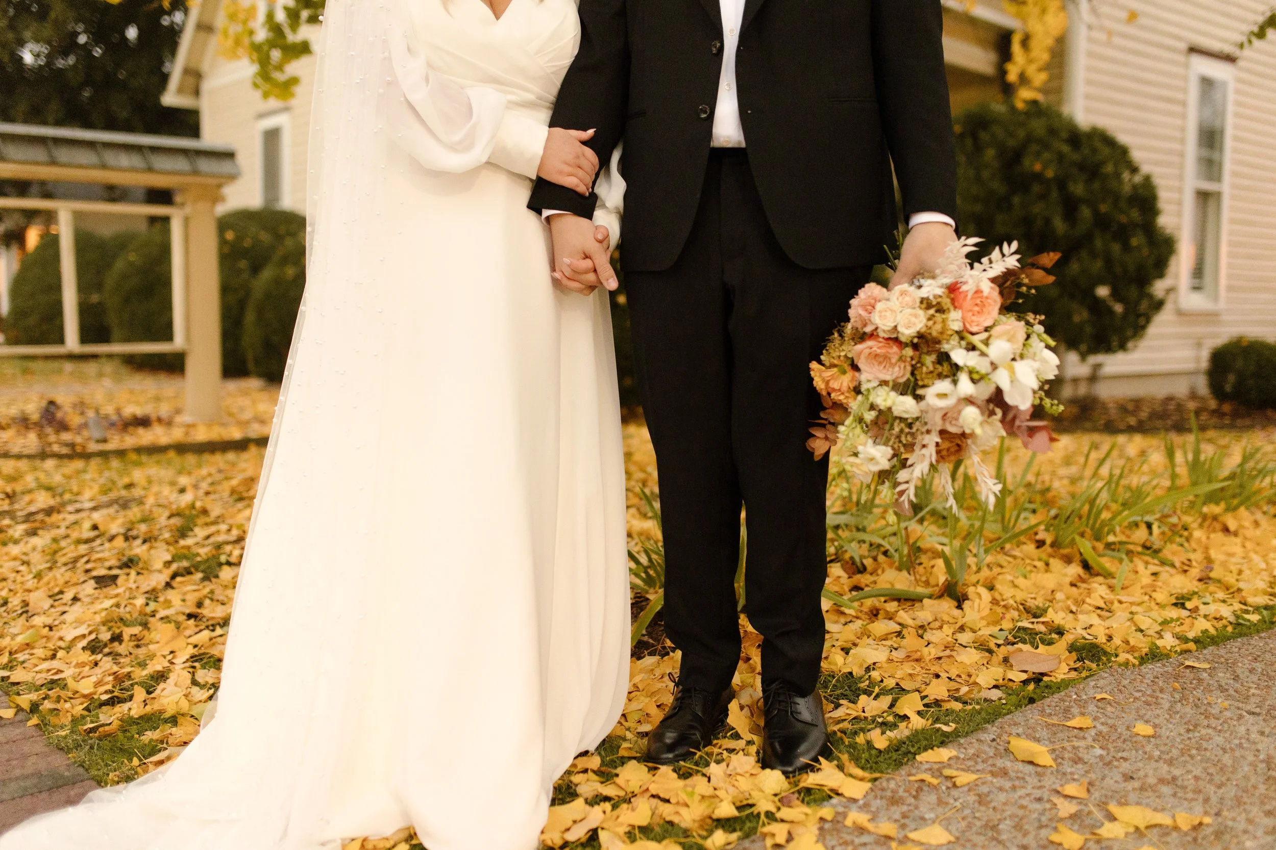 Close-up of newlyweds holding hands over golden autumn leaves during their fall wedding portraits.