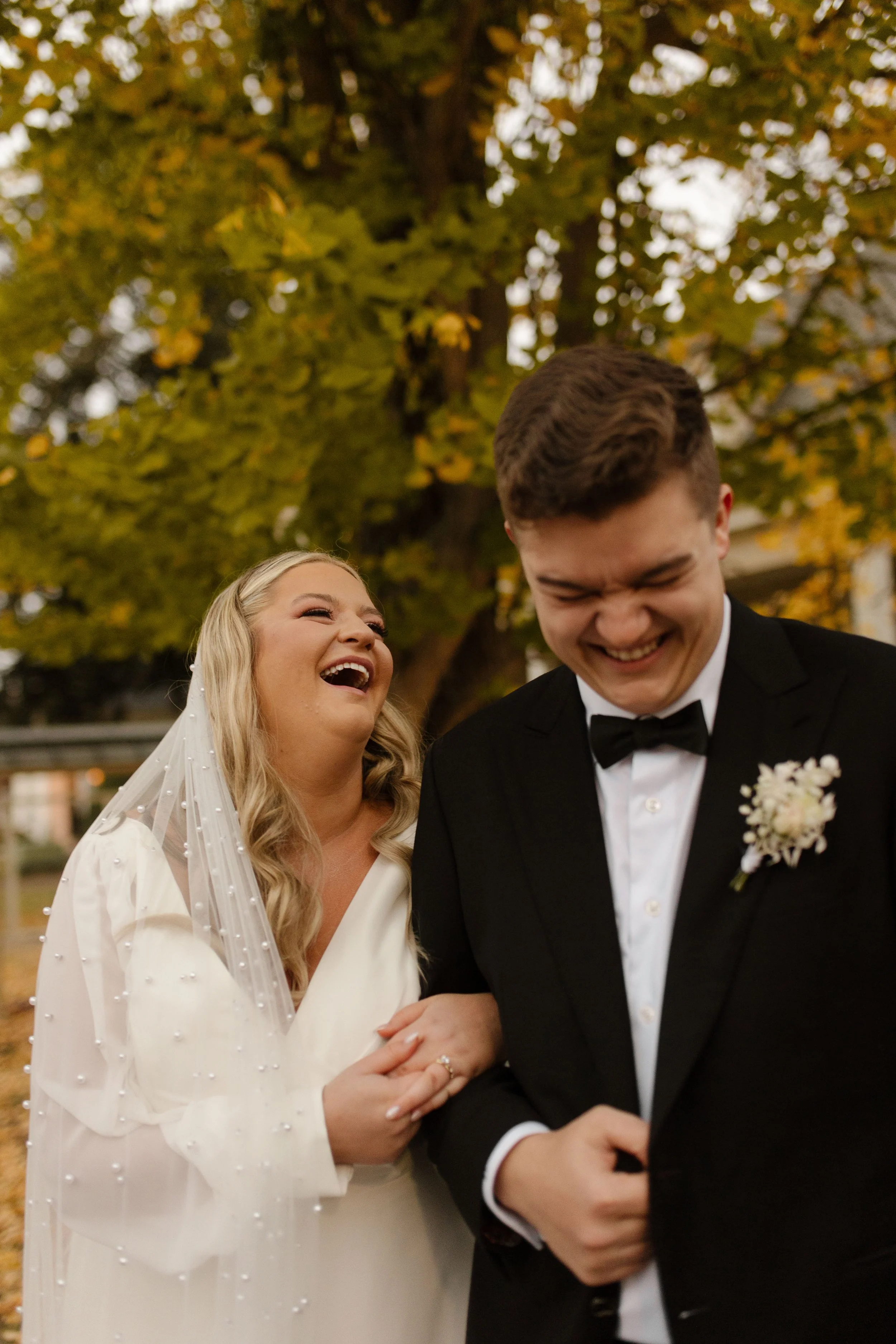 Bride and groom laughing together beneath golden autumn trees during their fall wedding portraits.