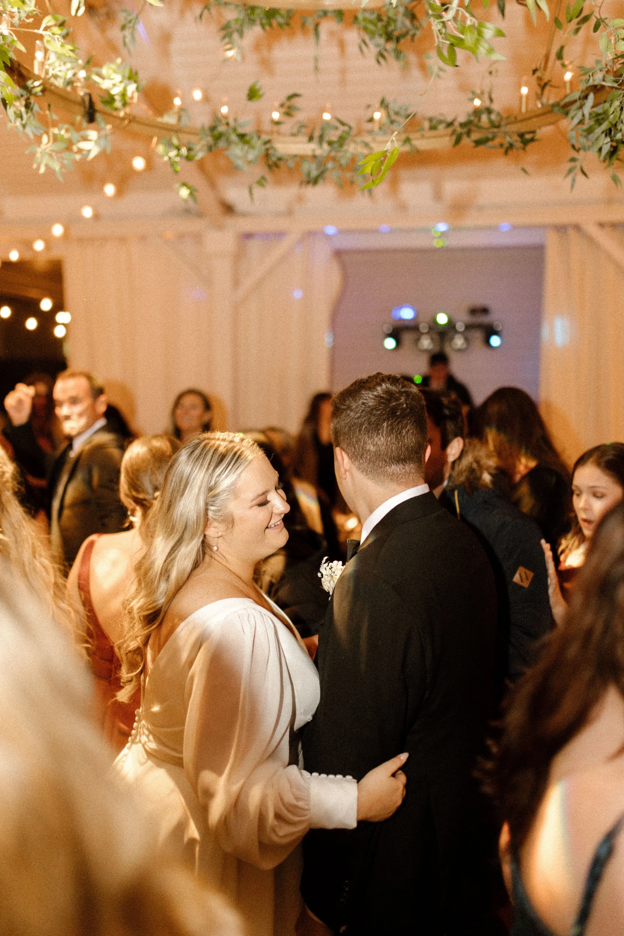 Bride and groom together on a crowded dance floor during their fall wedding reception beneath string lights and greenery.