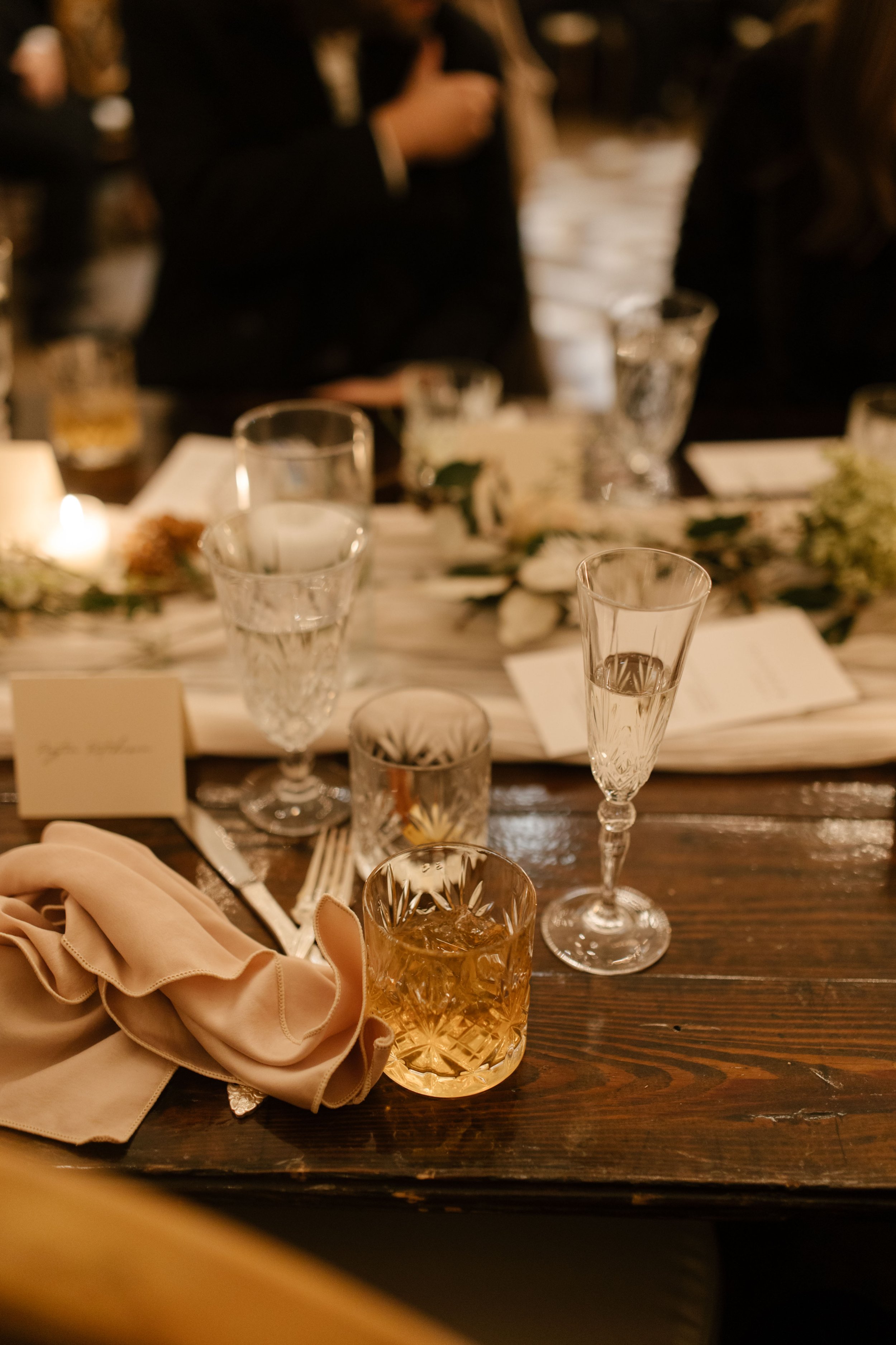 Champagne glass and cocktail on a warmly lit reception table with blush linens and greenery at a fall wedding.