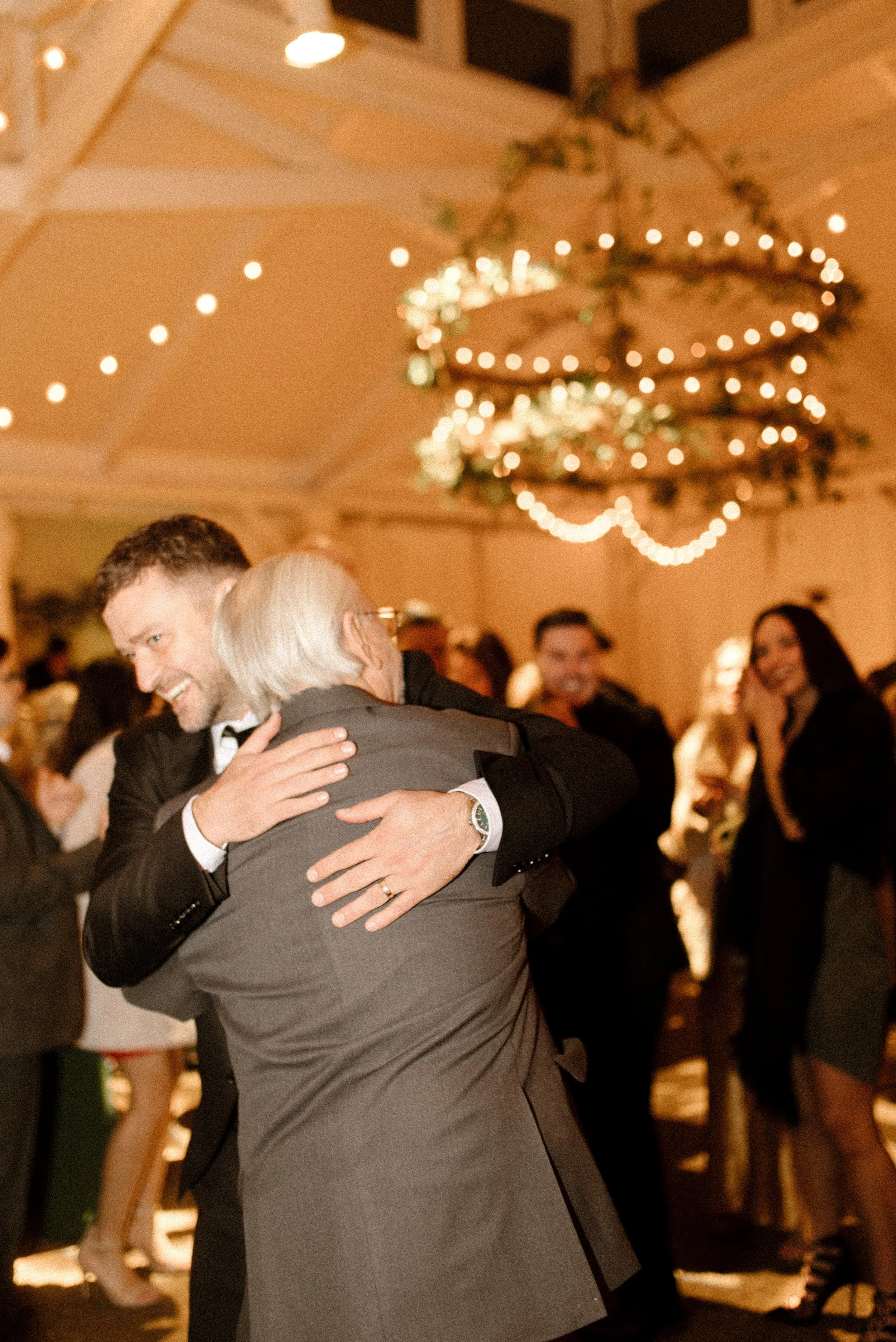 Candid reception photo of a wedding guest hugging an older family member beneath glowing string lights.