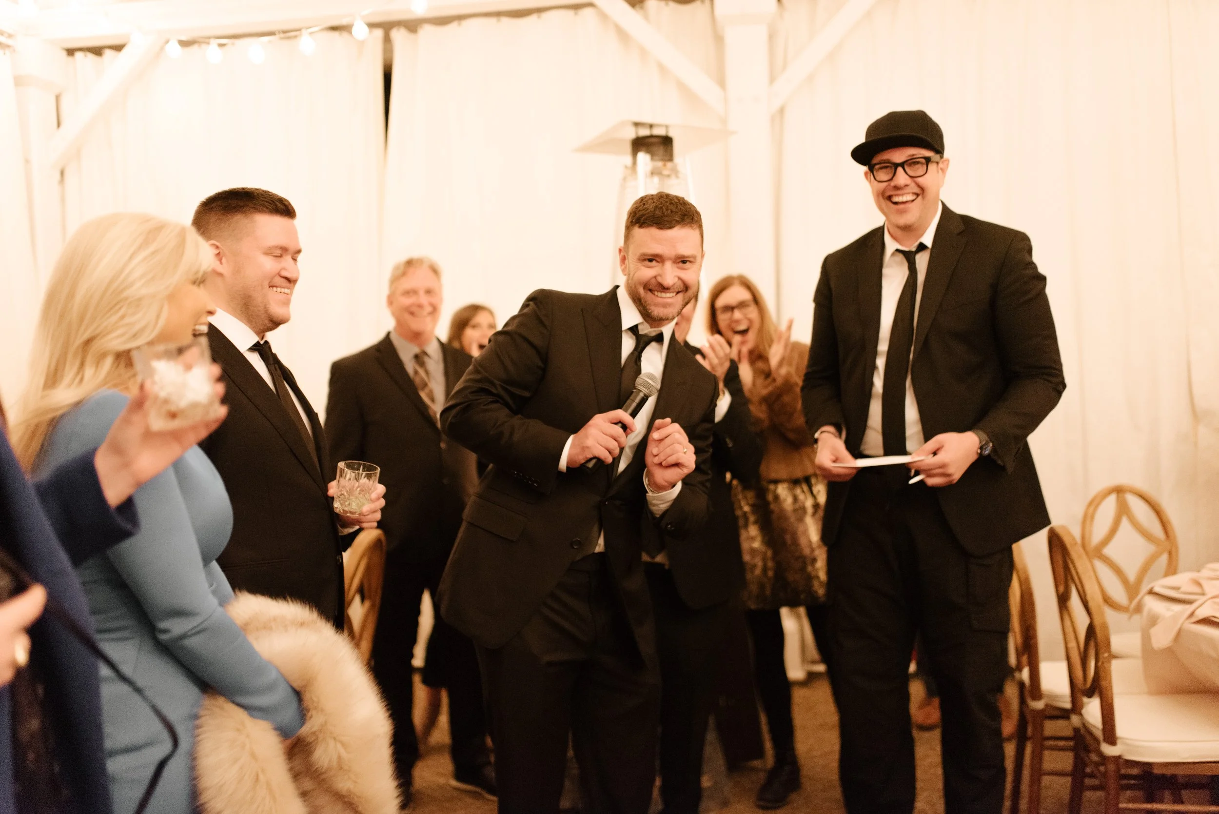 Wedding guests laughing during a lively reception toast beneath warm string lights at a fall wedding.
