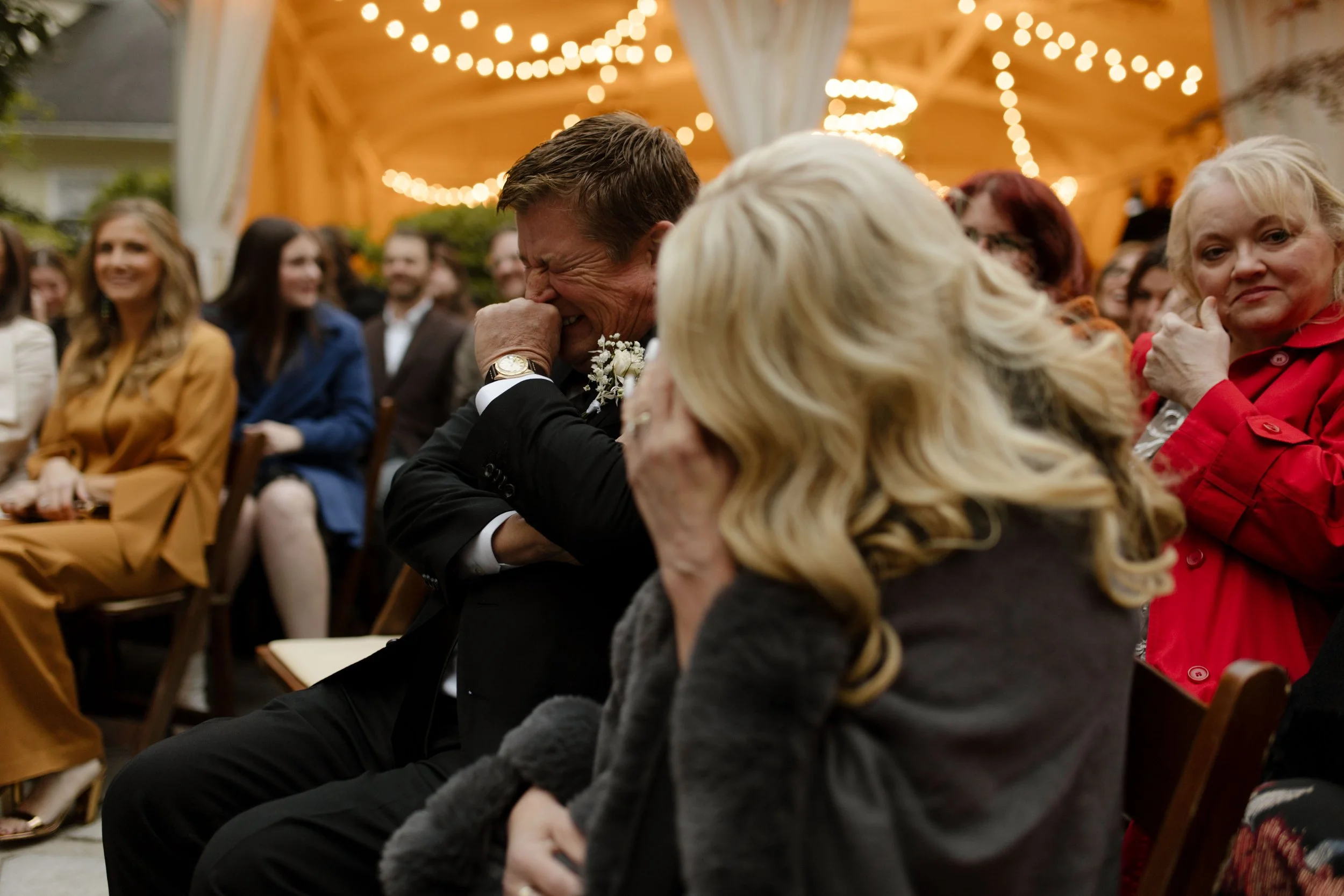 Emotional wedding guests wiping away tears while watching the couple exchange vows beneath glowing string lights.