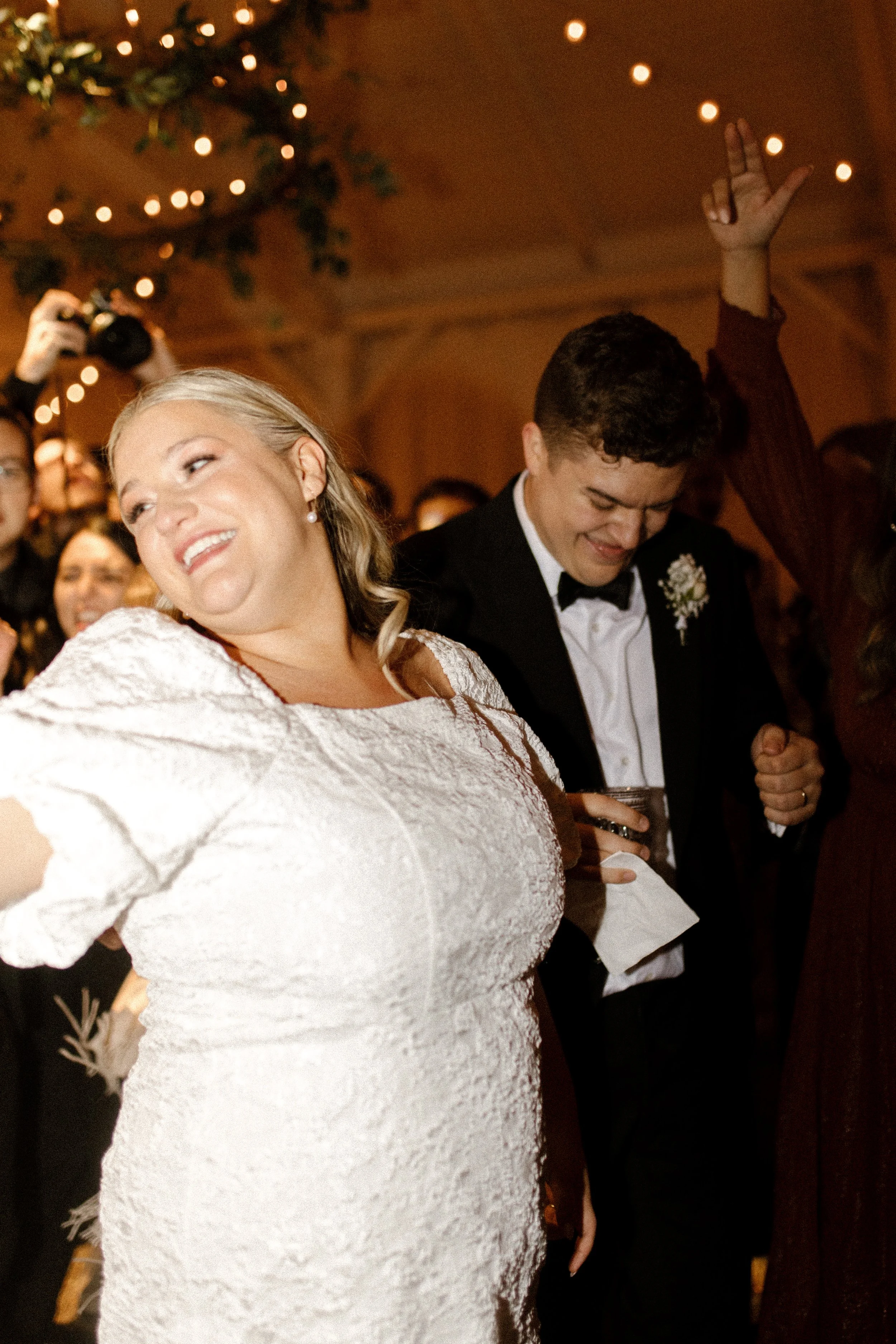 Bride and groom dancing with friends beneath warm string lights at their fall wedding reception.