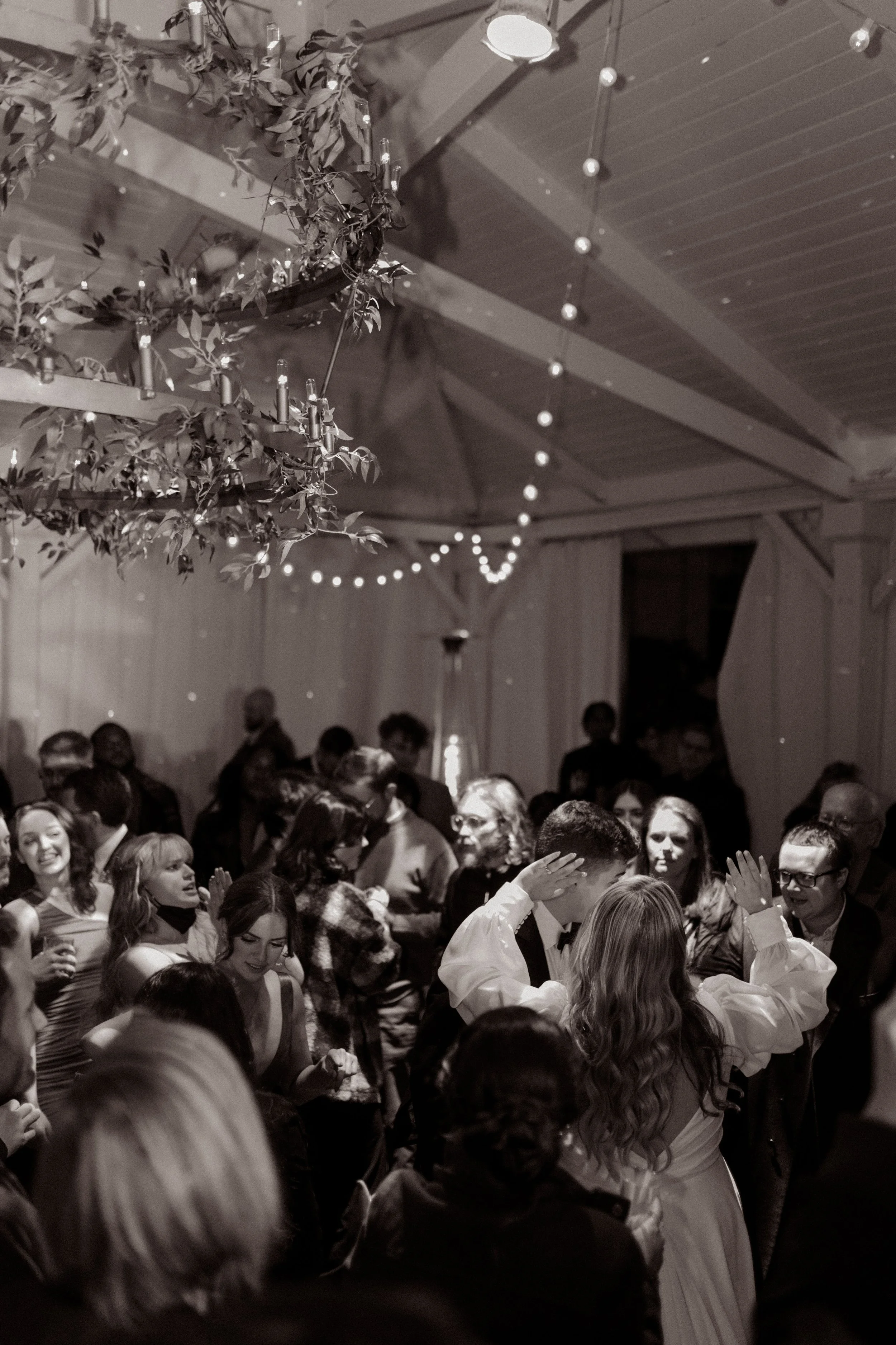 Black and white documentary wedding photo of a packed dance floor beneath hanging greenery and string lights.