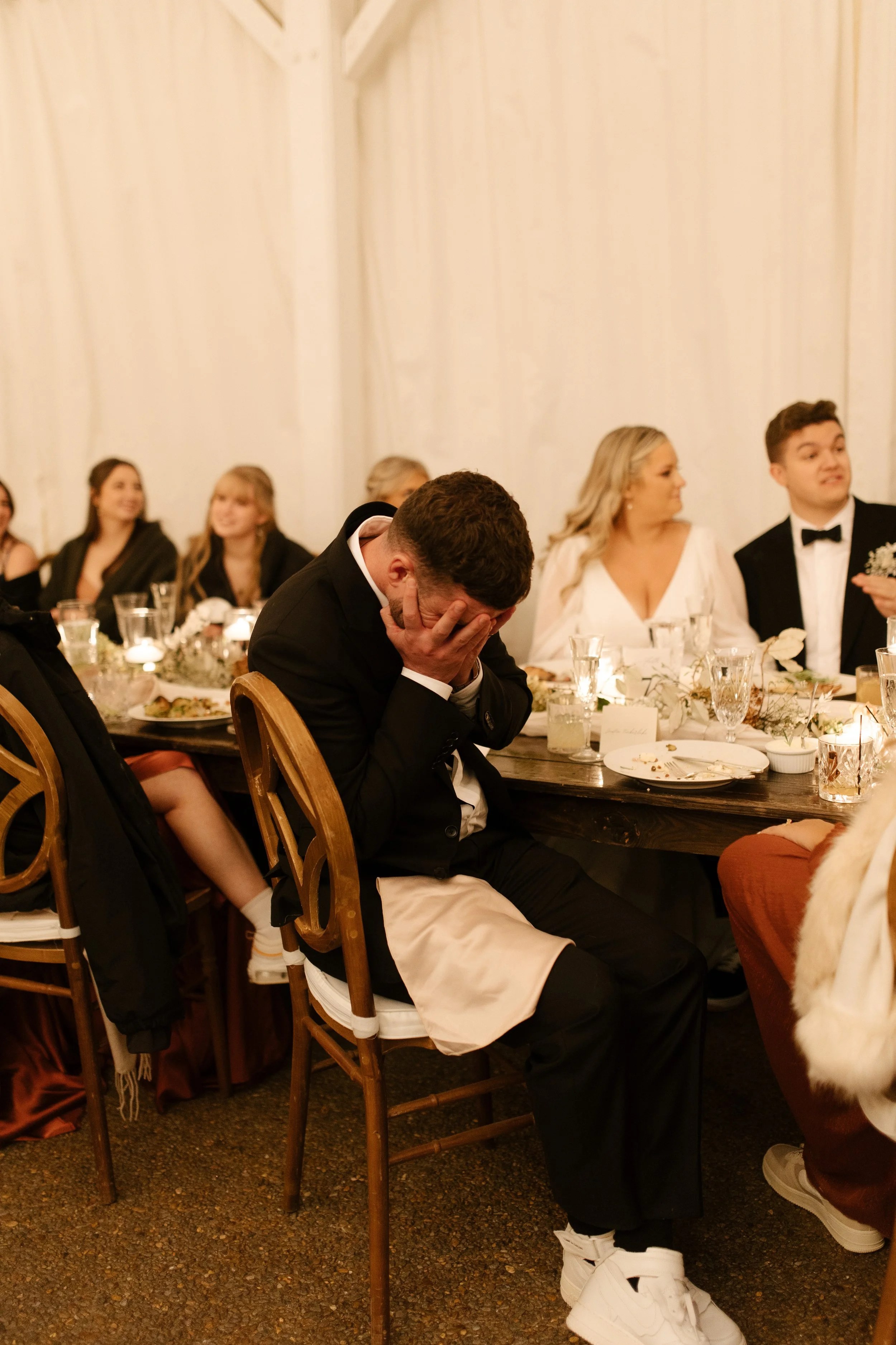 Emotional wedding guest covering his face during a reception speech as the newlyweds look on.
