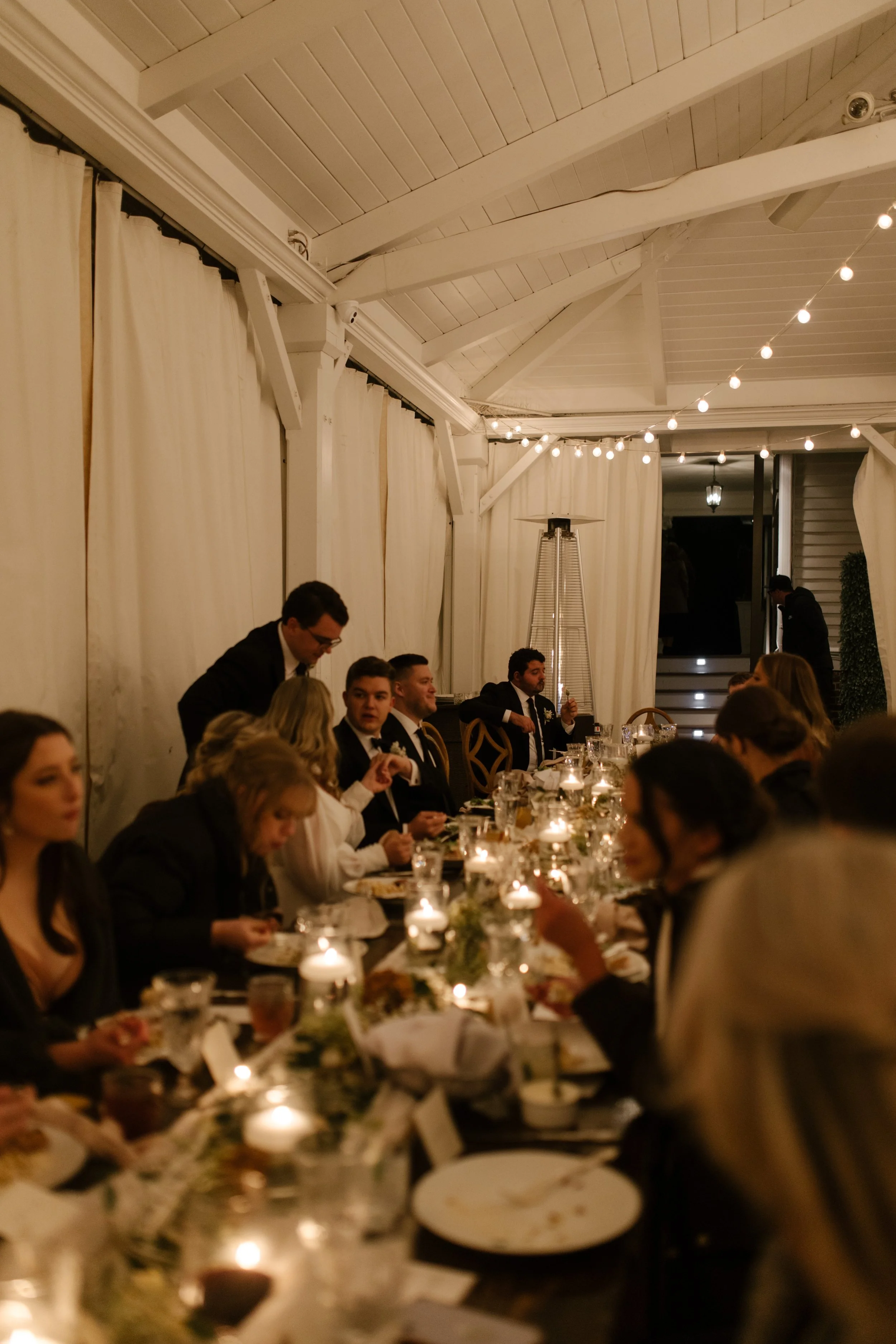 Candlelit wedding reception dinner with guests gathered around long tables beneath warm string lights.