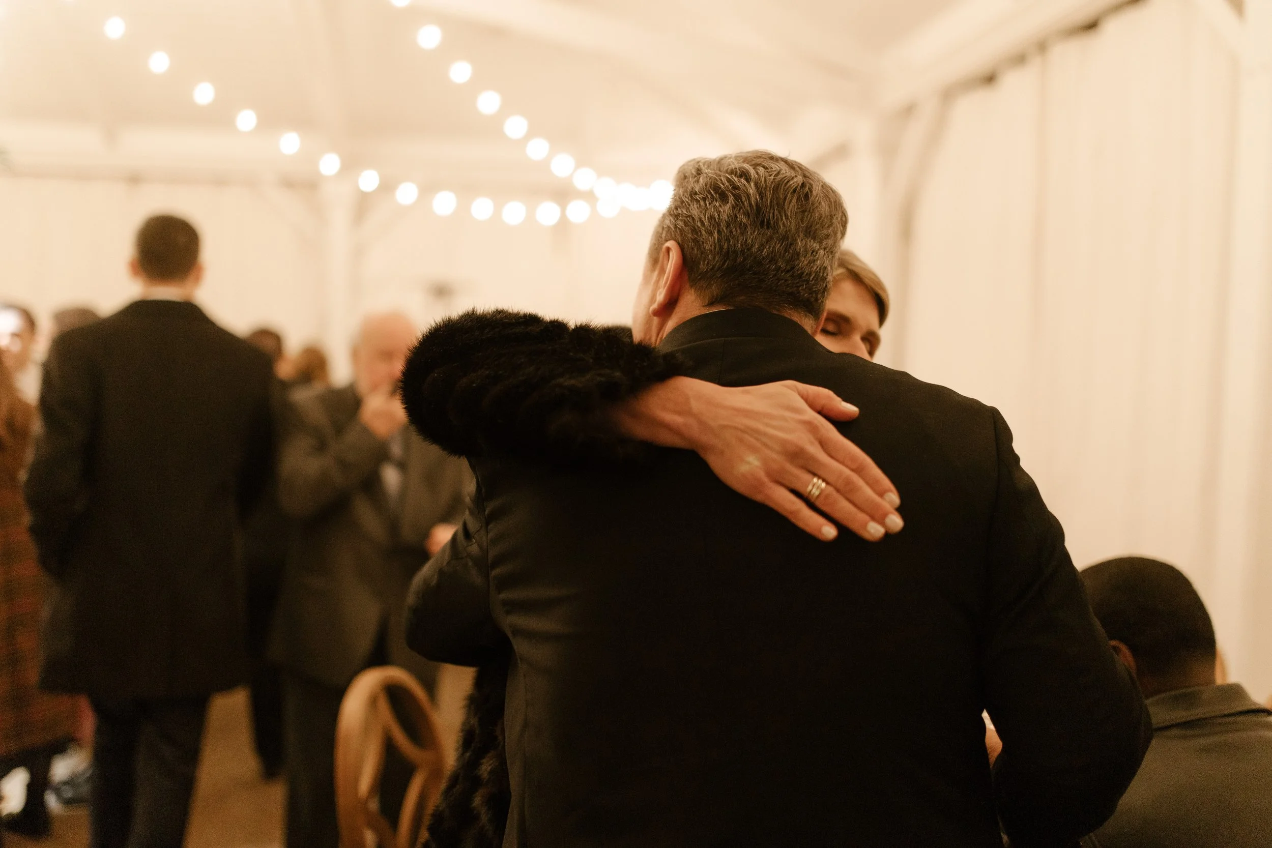 Candid wedding reception photo of two guests hugging beneath glowing string lights.