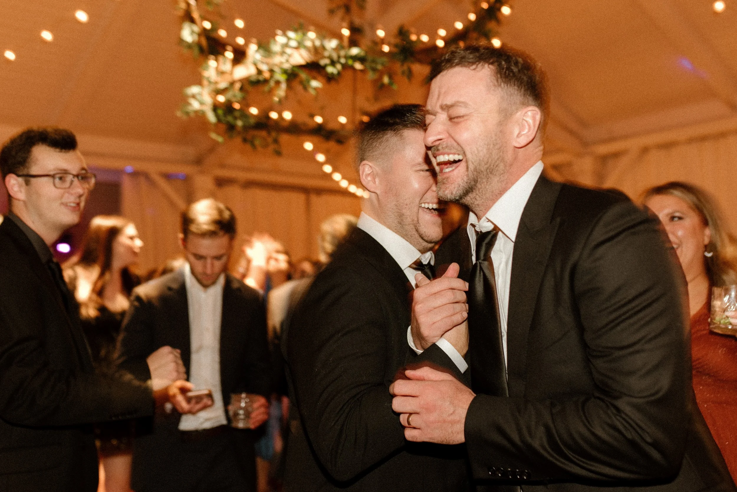 Wedding guests laughing and dancing together beneath warm string lights at the reception.