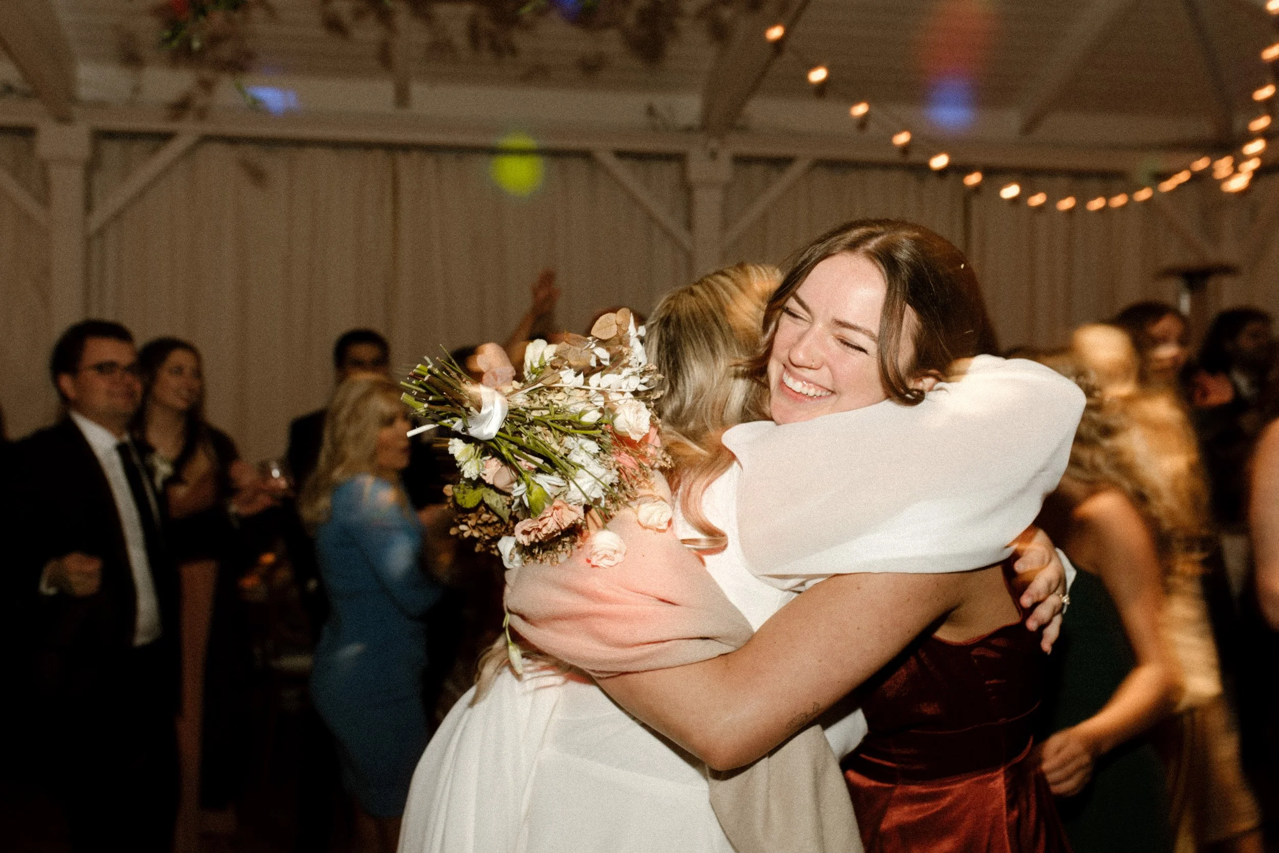 Bride hugging a smiling wedding guest while holding her bouquet during a lively fall wedding reception.