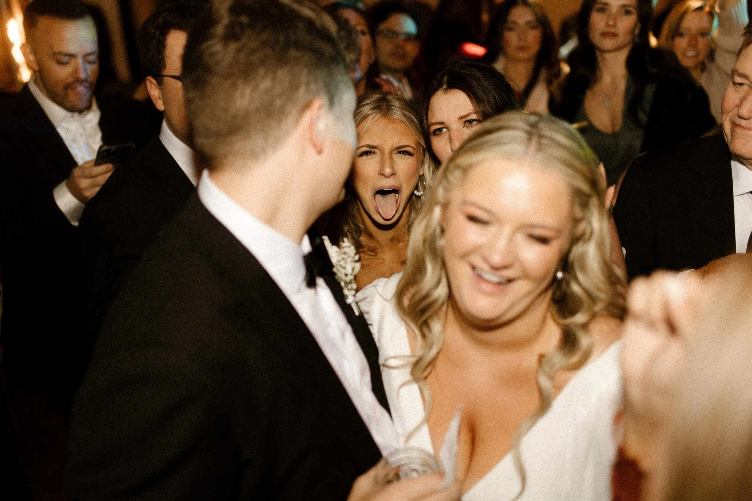 Bride dancing with the groom and friends on a packed wedding reception dance floor.