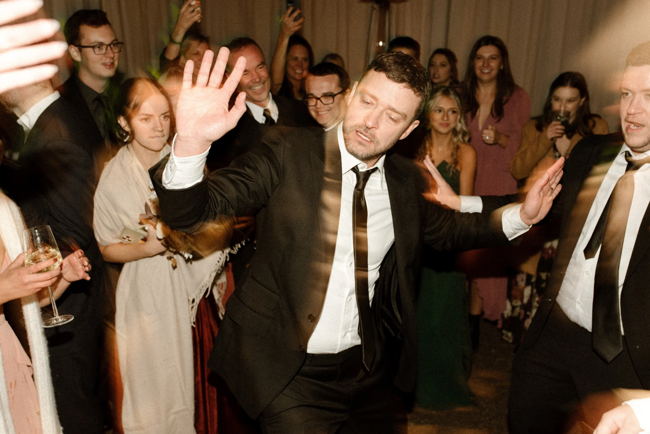 Wedding guest in a black suit dancing enthusiastically on a crowded reception dance floor.