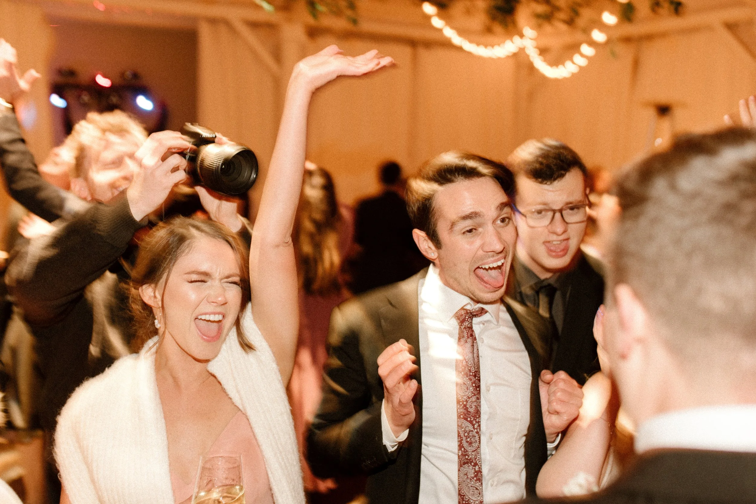 Guests cheering and dancing beneath warm string lights during a lively fall wedding reception.