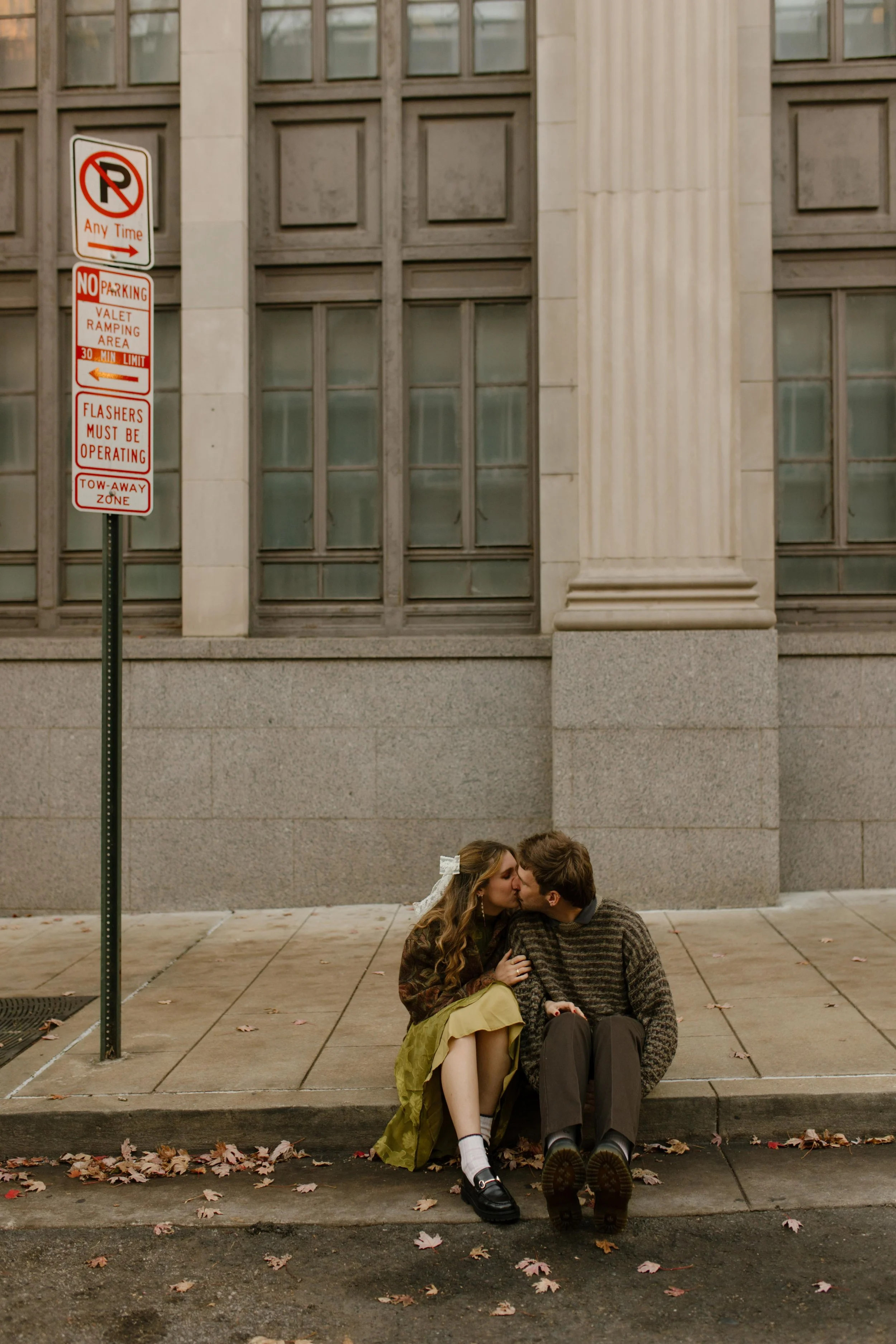 Couple kissing while seated on a city curb during engagement photos in NYC, surrounded by fallen autumn leaves.