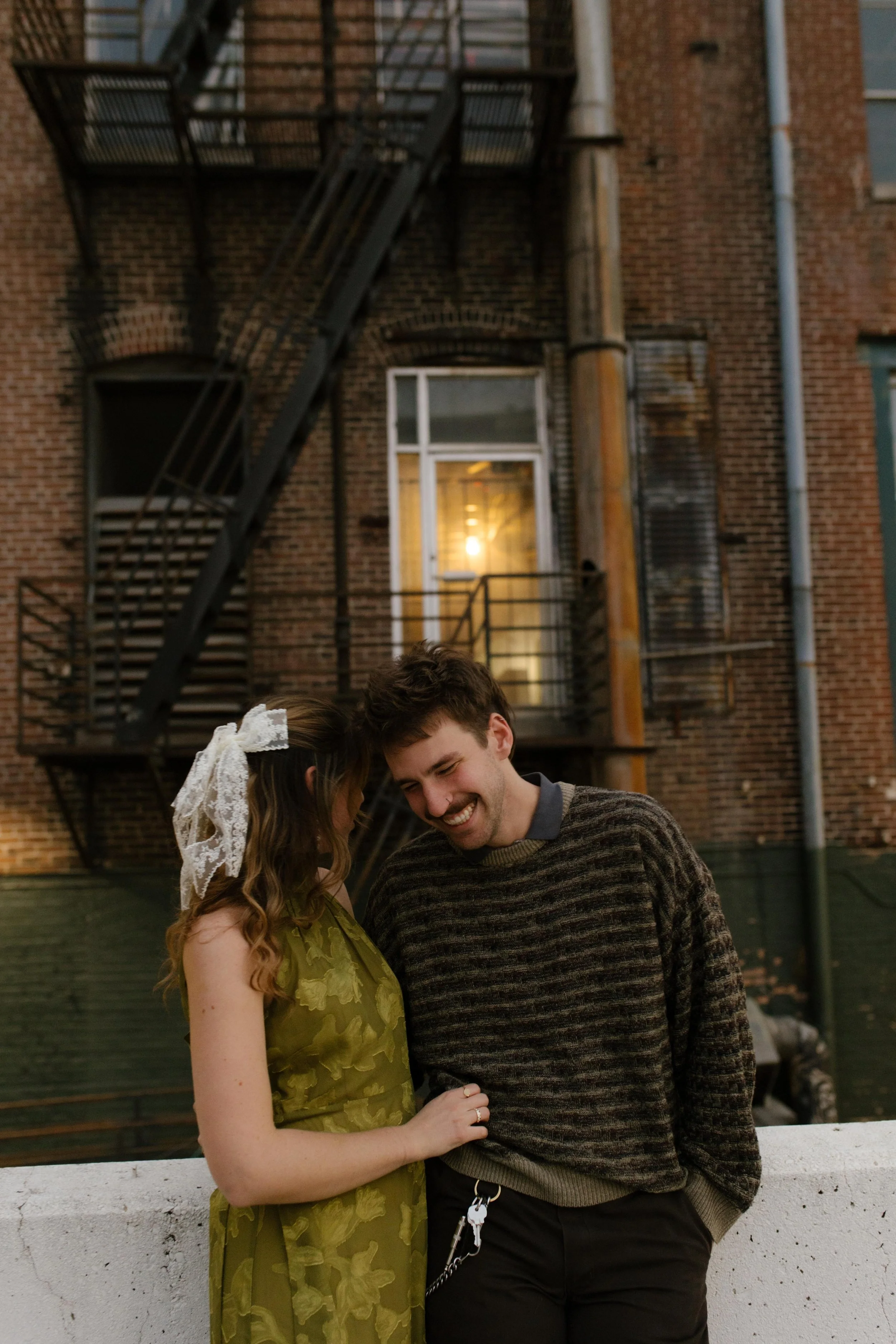 Engaged couple laughing together in front of a brick building and fire escape during a candid city portrait.