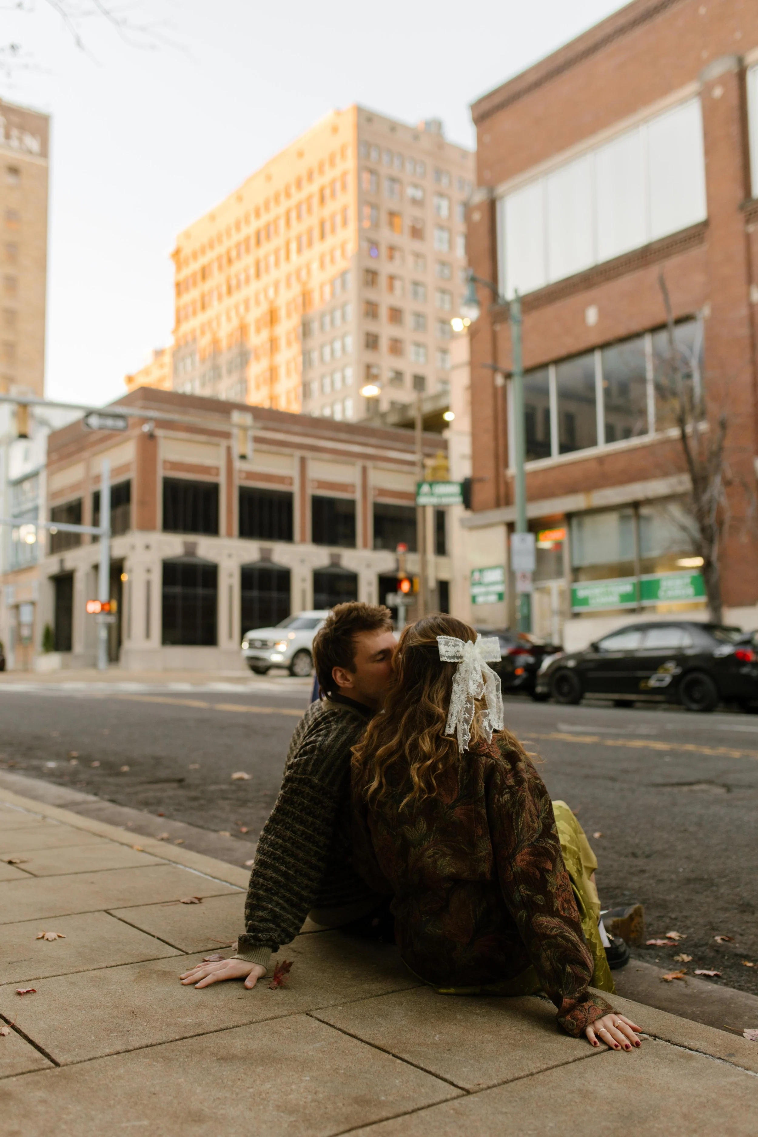 Engaged couple kissing on a downtown sidewalk with tall buildings and city streets surrounding them.