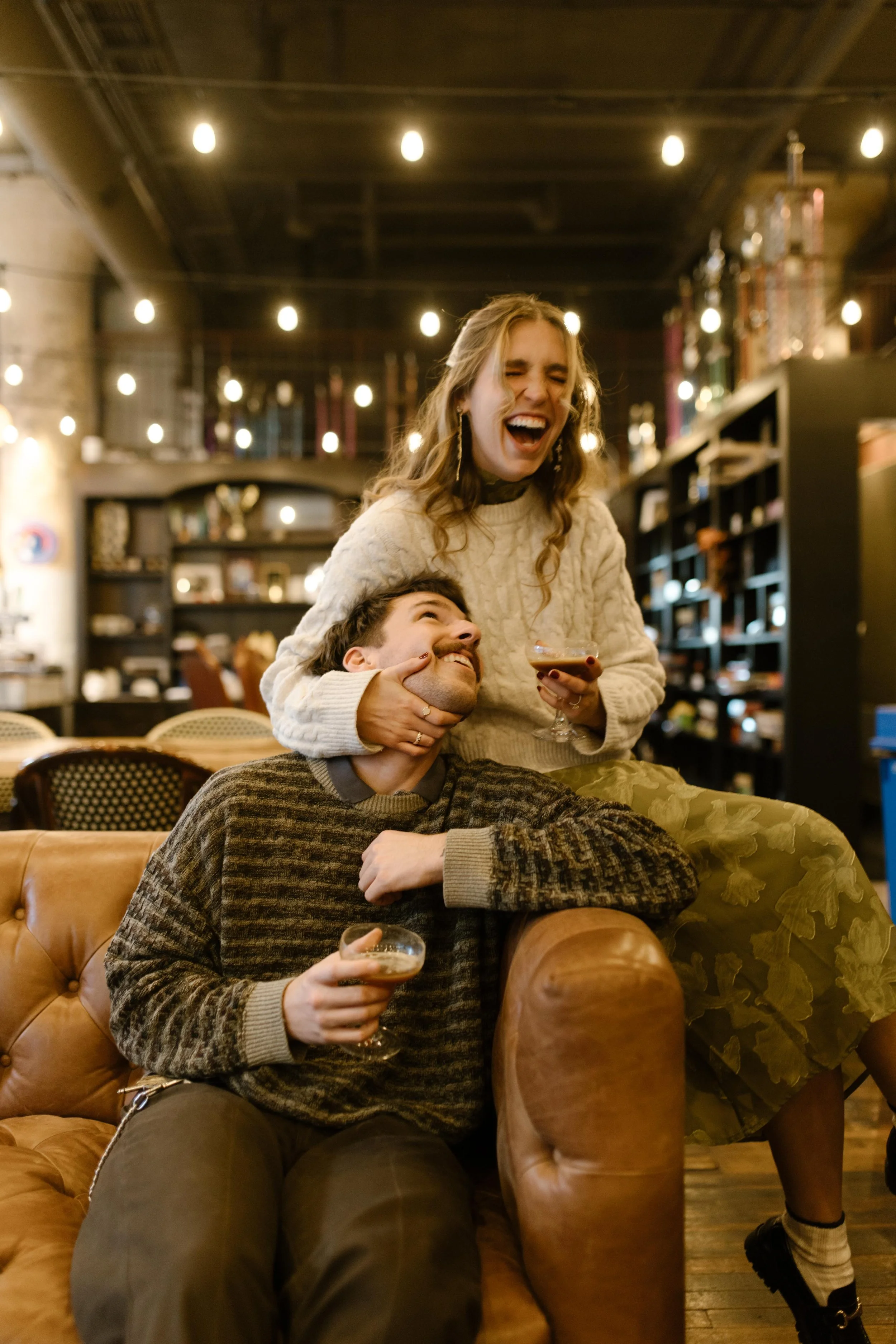 Couple laughing and sharing cocktails during engagement photos in NYC inside a cozy, warmly lit lounge.