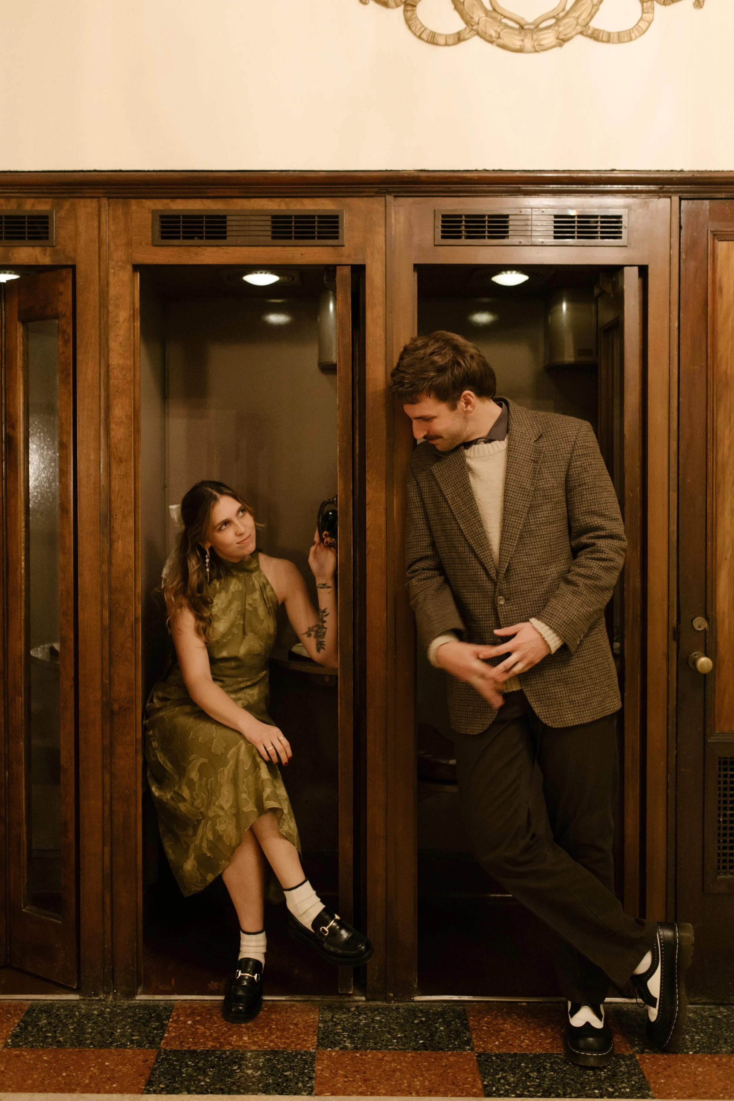Couple posing in neighboring vintage booths during engagement photos in NYC, wearing retro-inspired outfits.