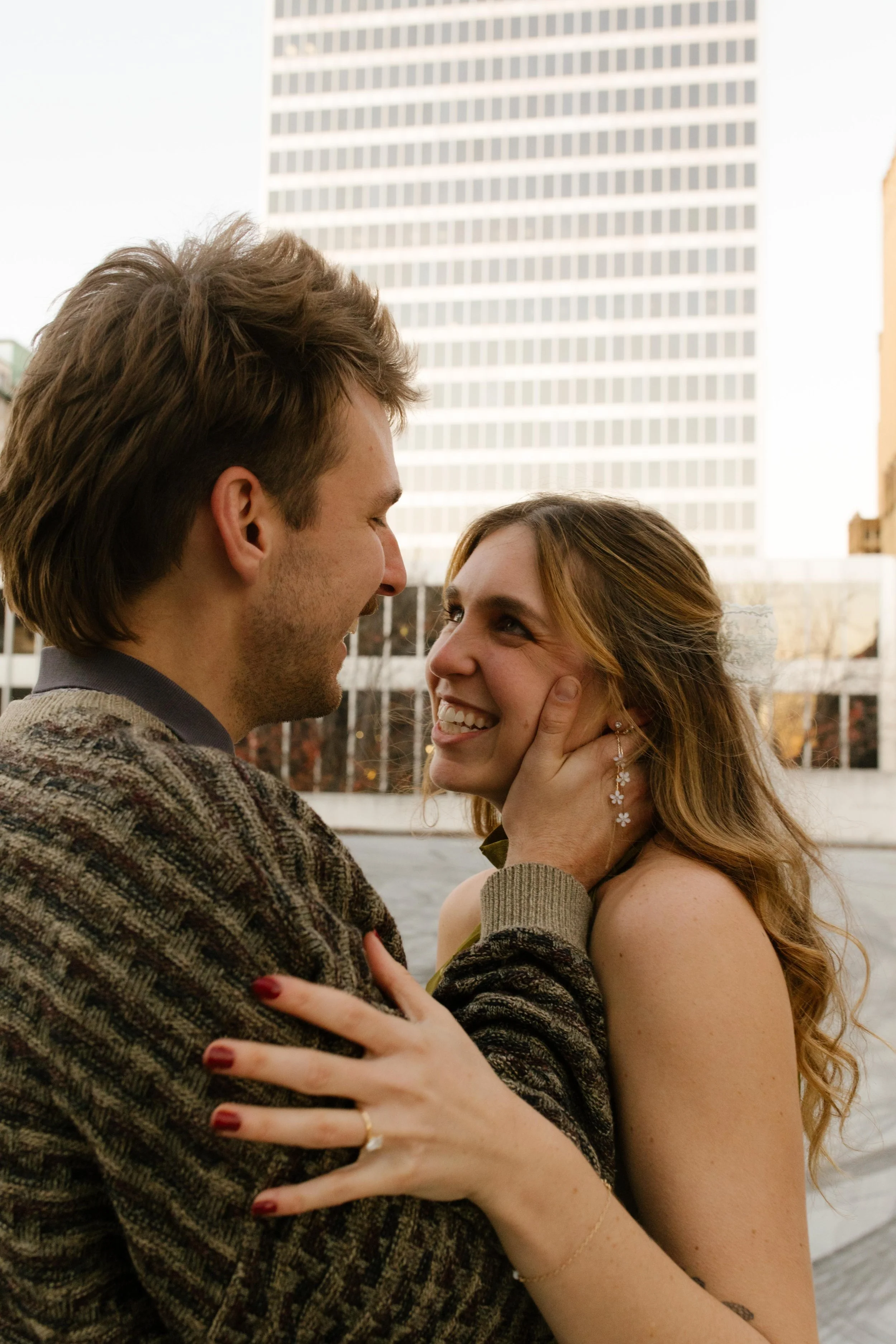 Close-up portrait of an engaged couple smiling at each other outdoors with city buildings in the background.