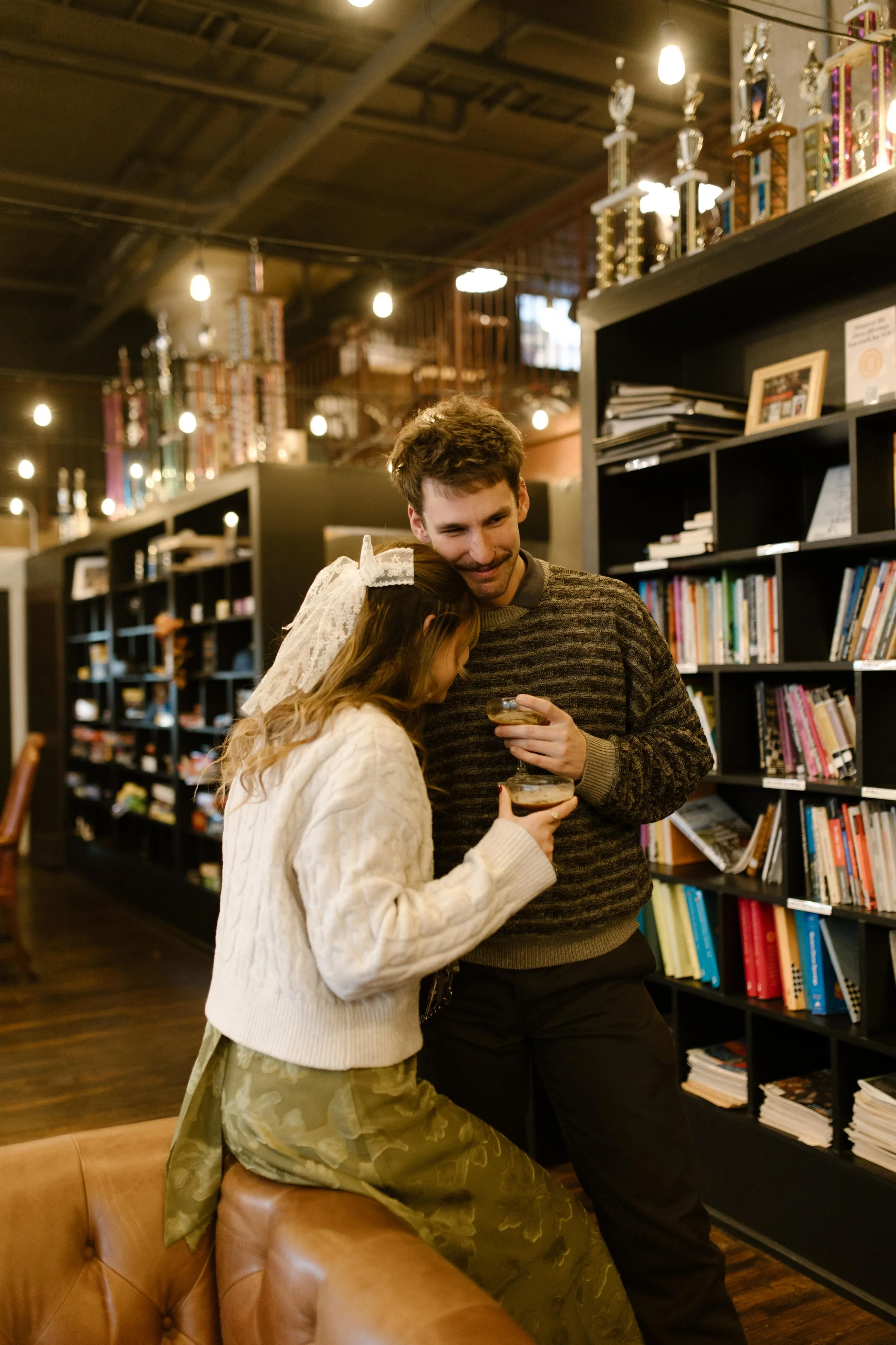 Couple sharing a cozy drink together during engagement photos in NYC inside a warmly lit lounge lined with books and trophies.