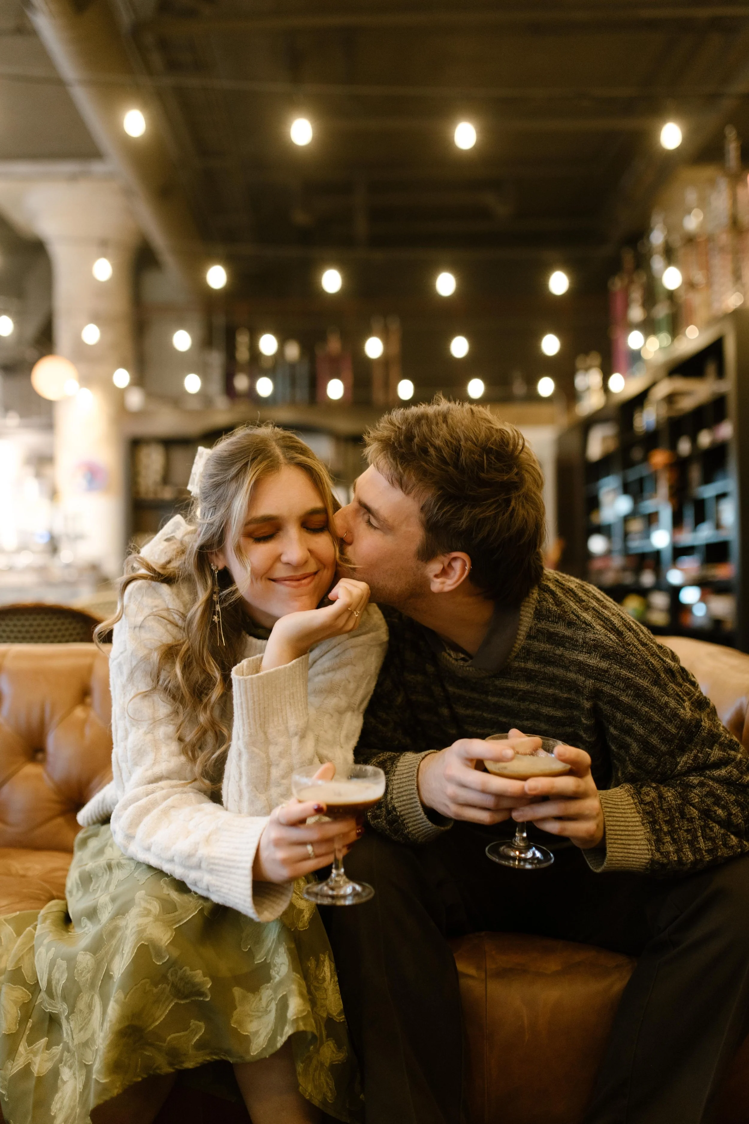 Engaged couple sharing cocktails on a leather couch as he kisses her cheek.