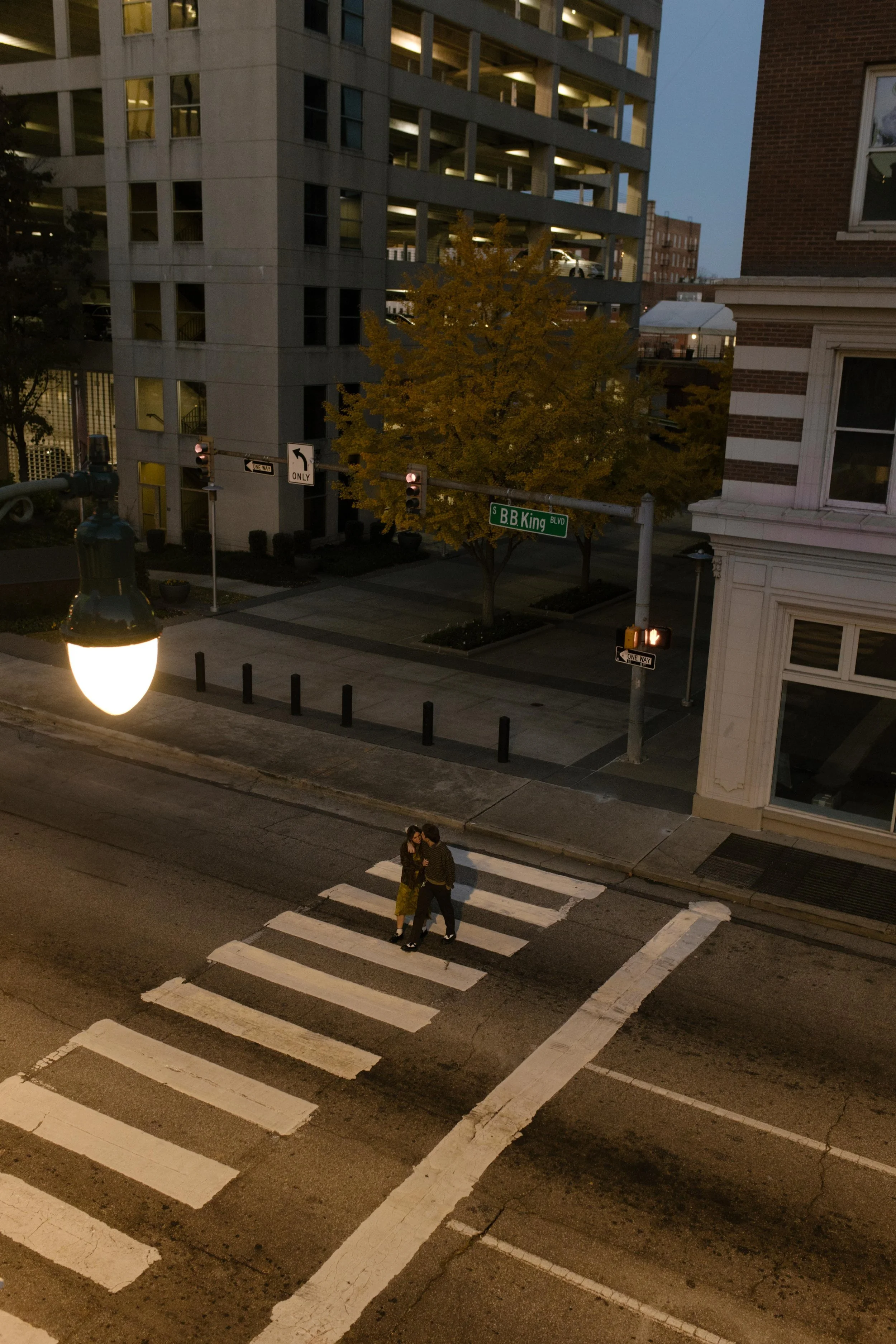 Couple standing together in a city crosswalk at dusk beneath glowing streetlights.