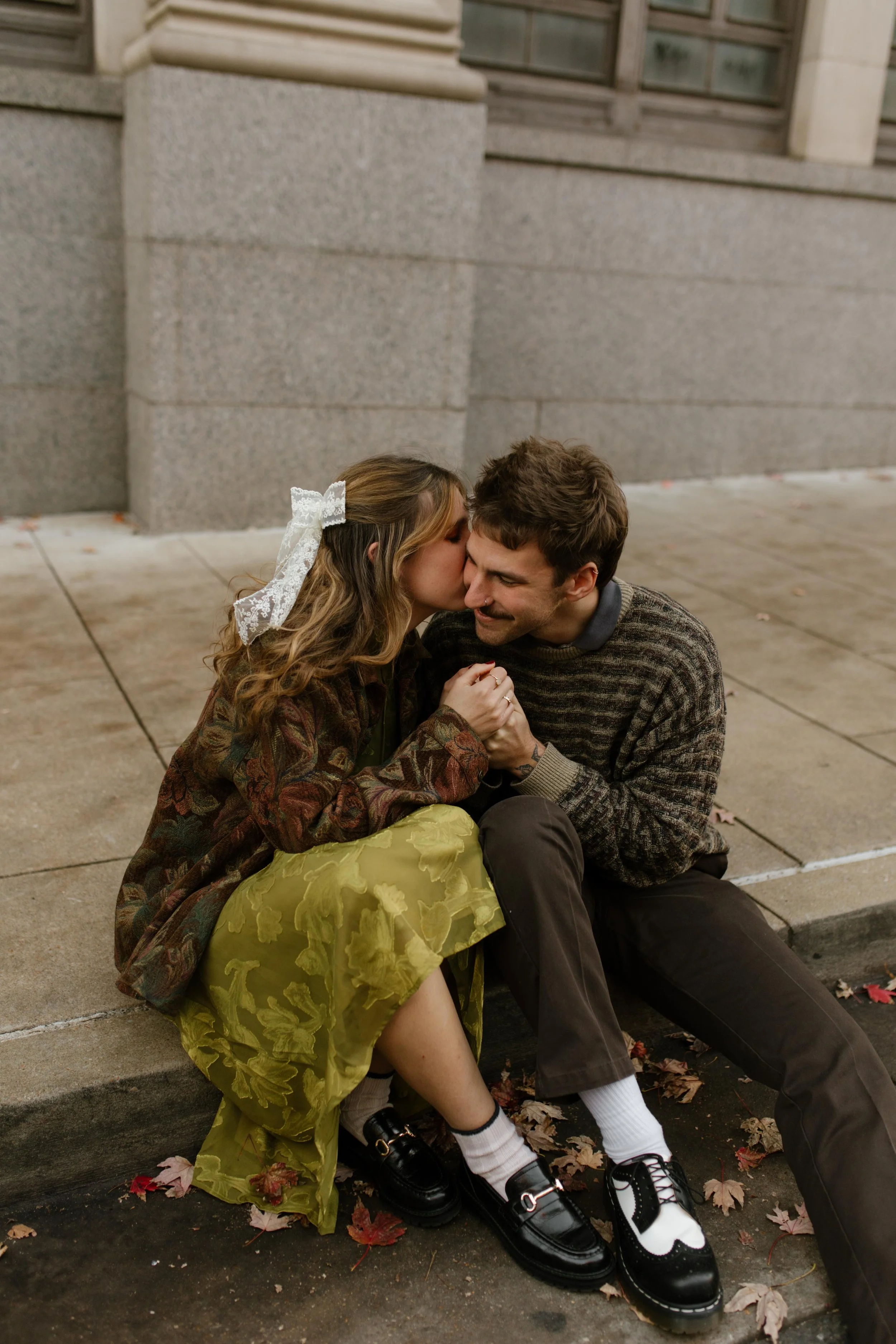 Engaged couple cuddling on a curb during engagement photos in NYC, with autumn leaves scattered around them.