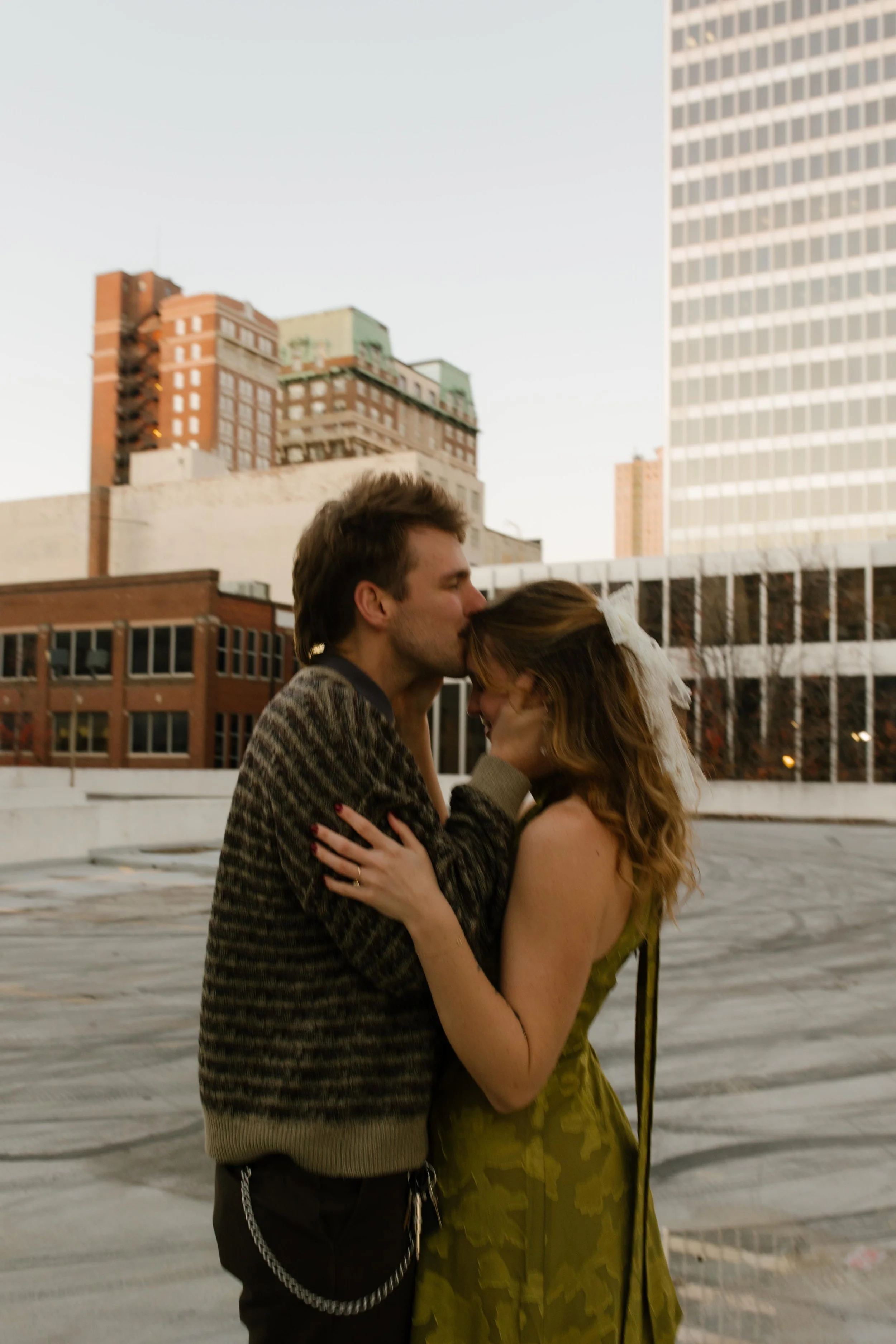 Couple embracing on a rooftop as he kisses her forehead with city buildings in the background.