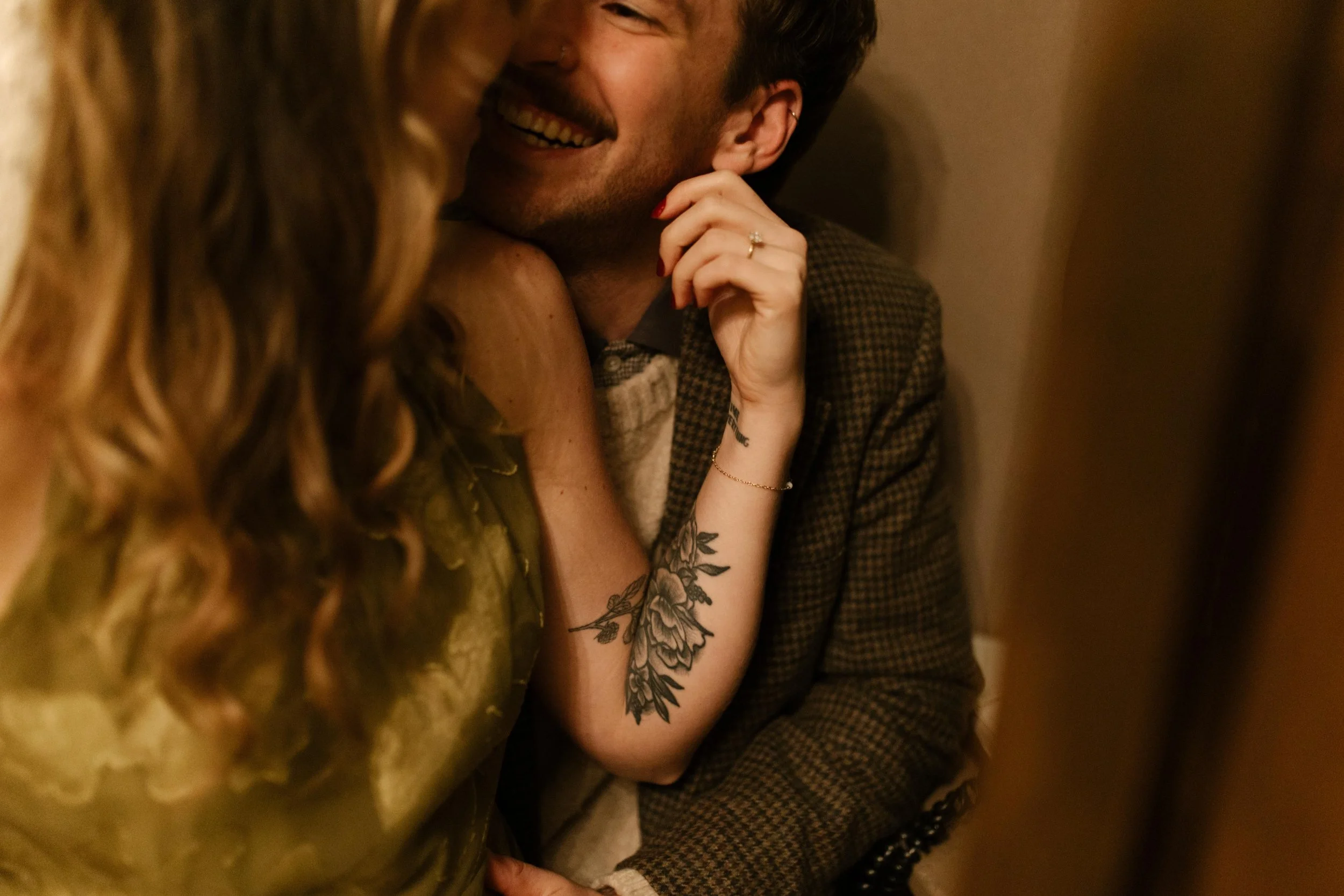 Candid close-up of an engaged couple laughing together, with her floral arm tattoo and engagement ring visible.