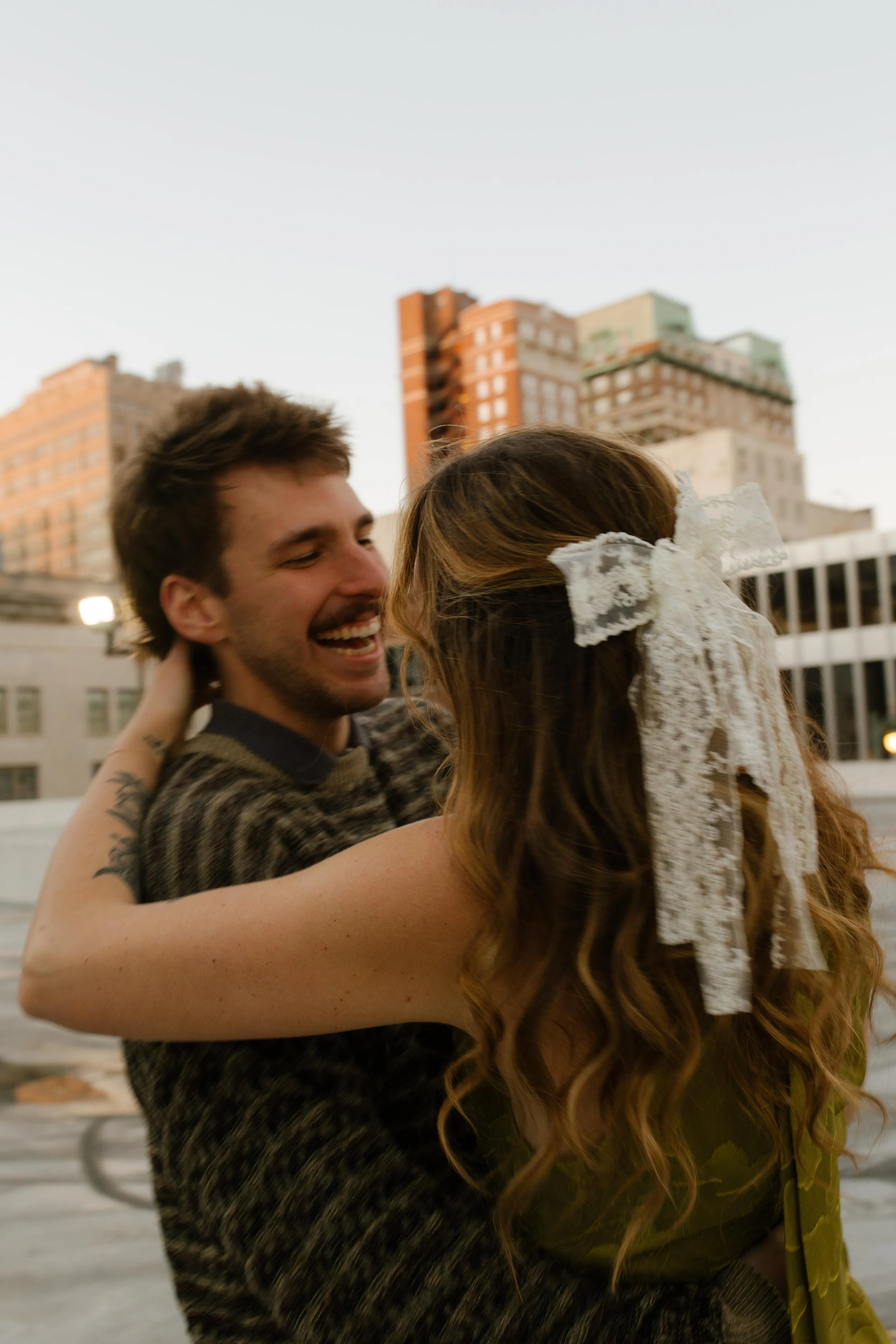 Couple laughing together on a rooftop during engagement photos in NYC, with a lace bow tied in her hair.