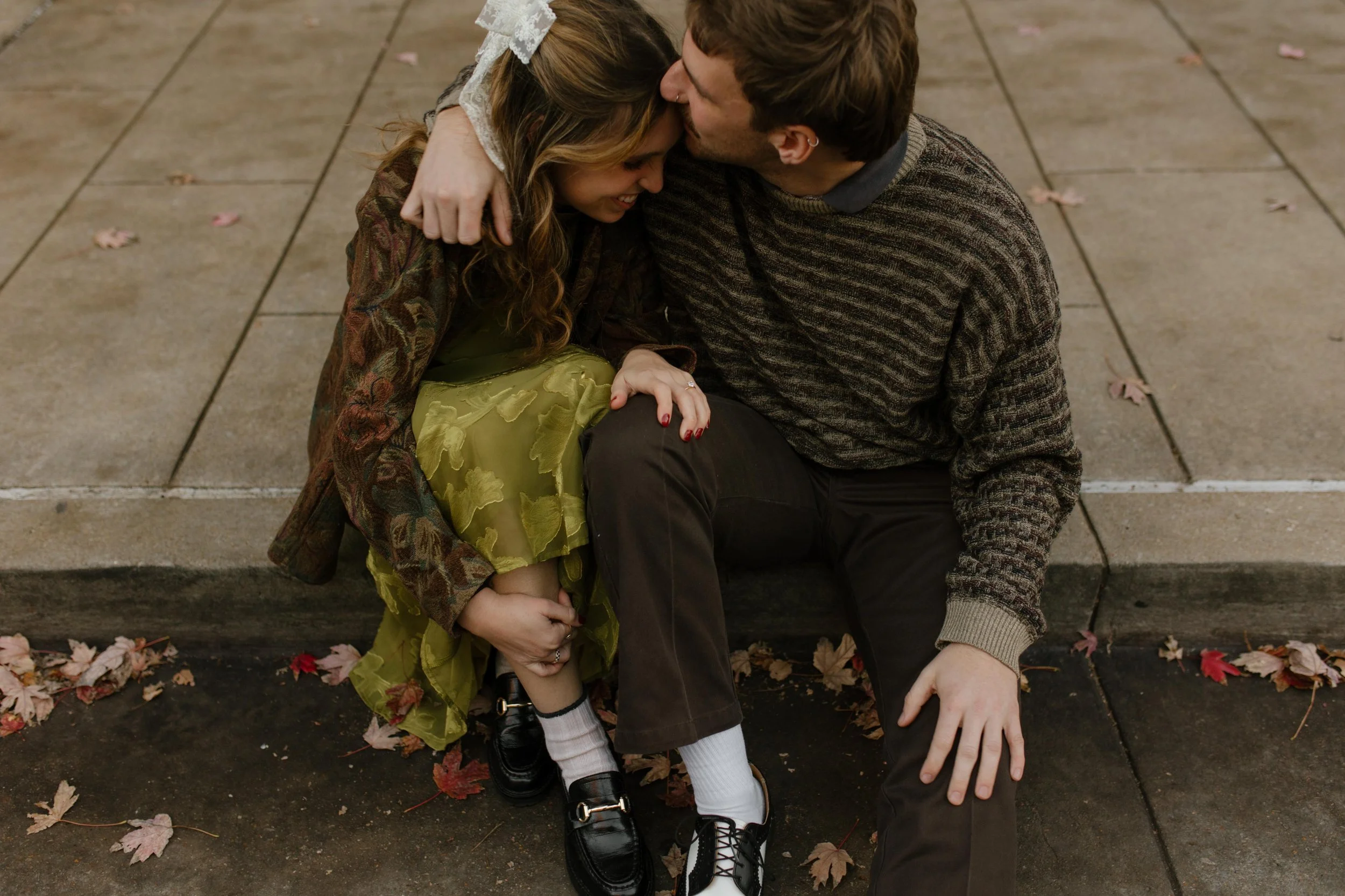 Engaged couple cuddling on a curb during engagement photos in NYC, with autumn leaves scattered around their feet.