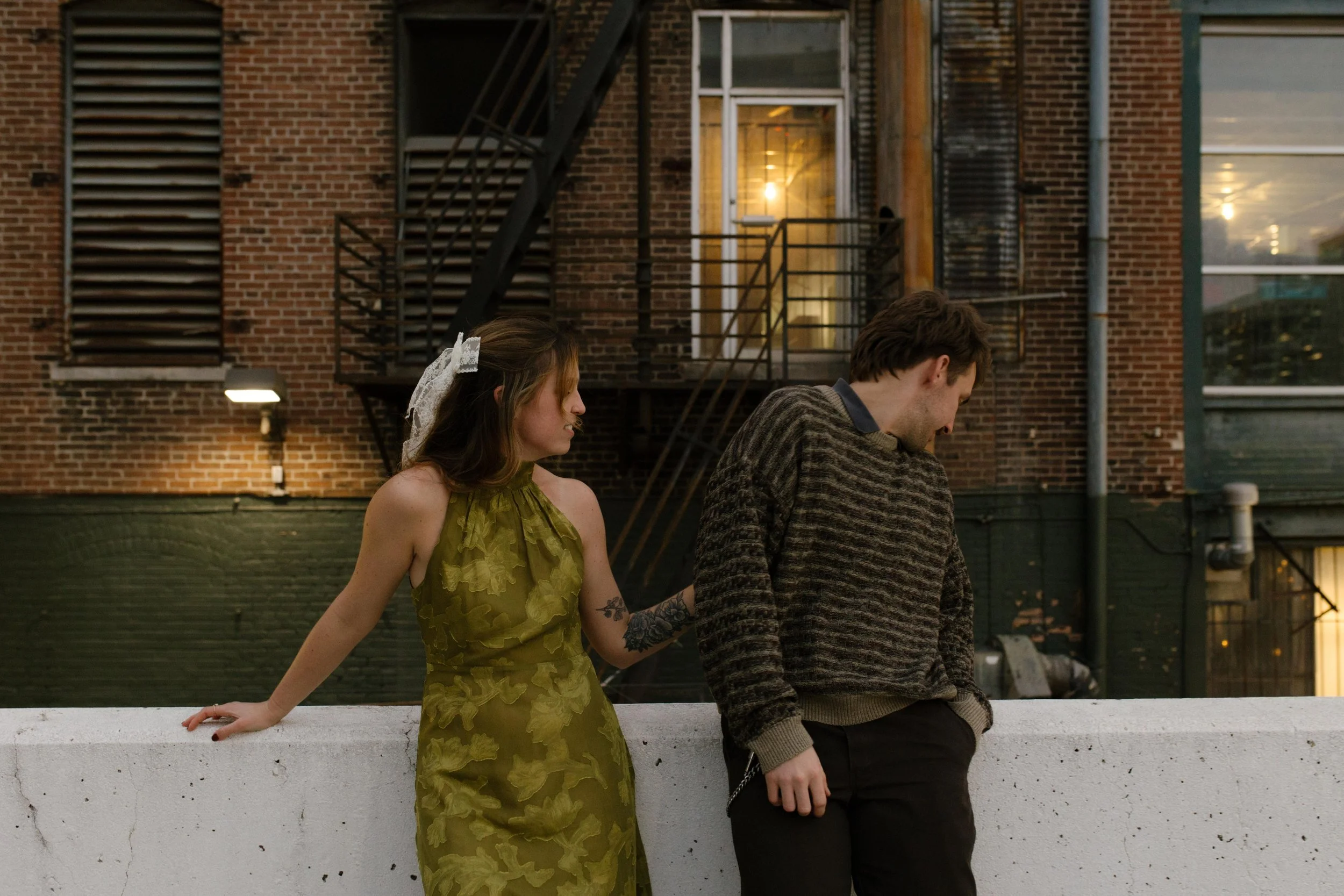 Engaged couple posing against a rooftop wall with brick buildings and fire escapes behind them.