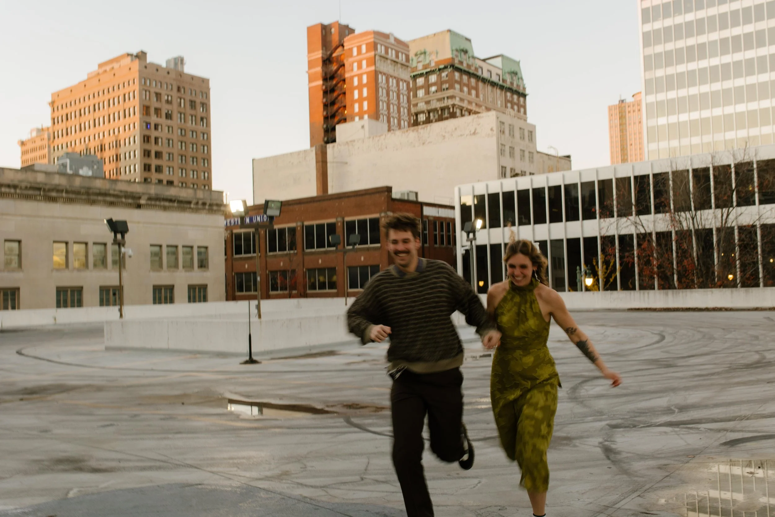 Engaged couple running hand in hand across a rooftop with downtown buildings in the background.