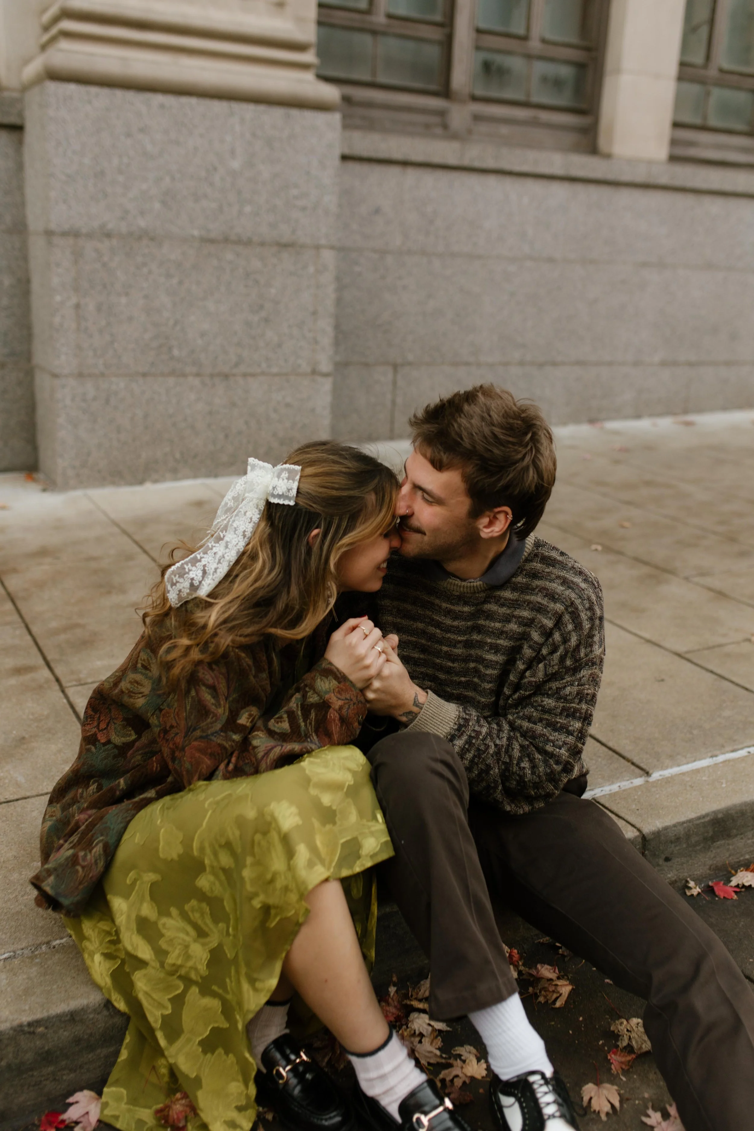Engaged couple cuddling together on a city curb surrounded by fallen autumn leaves.