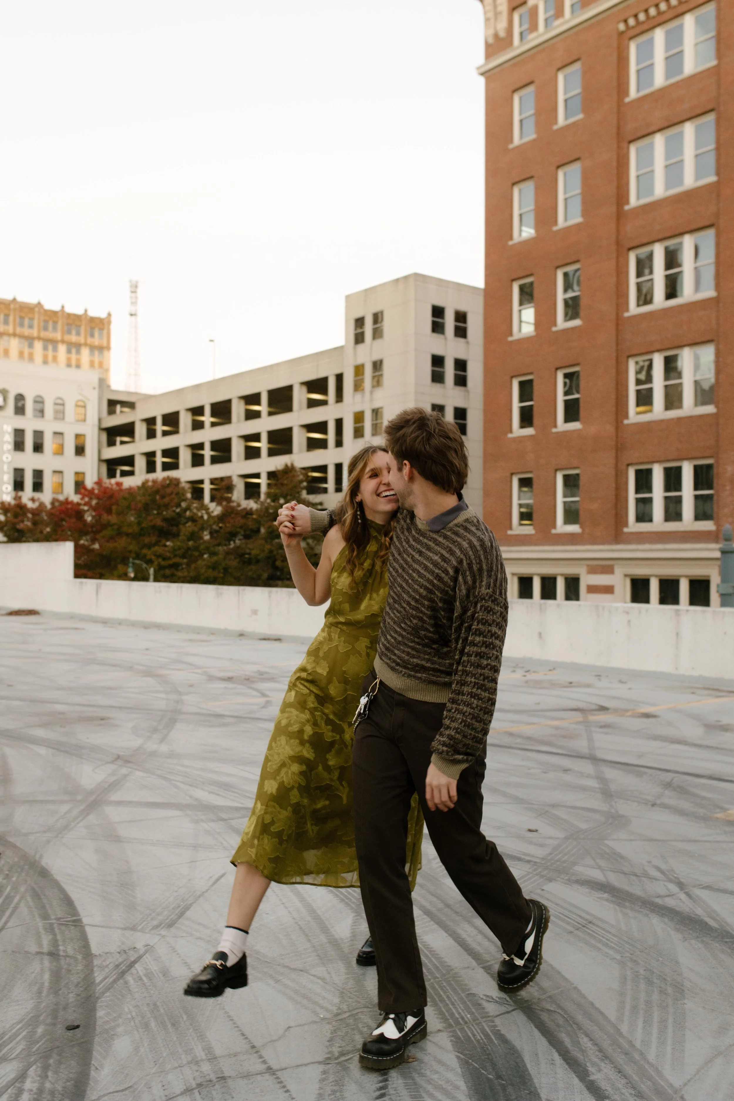 Engaged couple walking hand in hand across a rooftop with downtown buildings and autumn trees behind them.