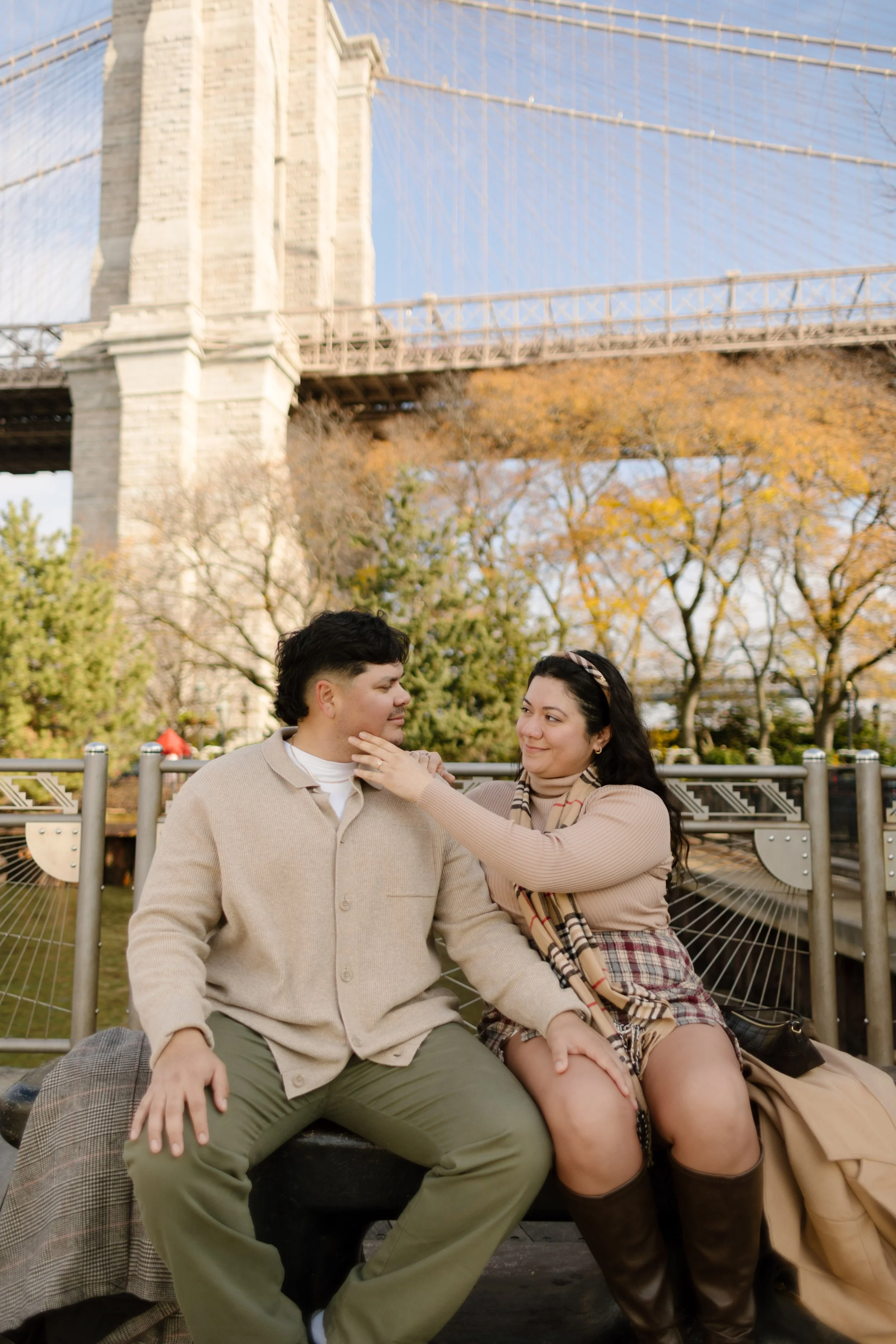 Cozy fall engagement portrait of a couple sitting together near the Brooklyn Bridge in neutral autumn outfits.