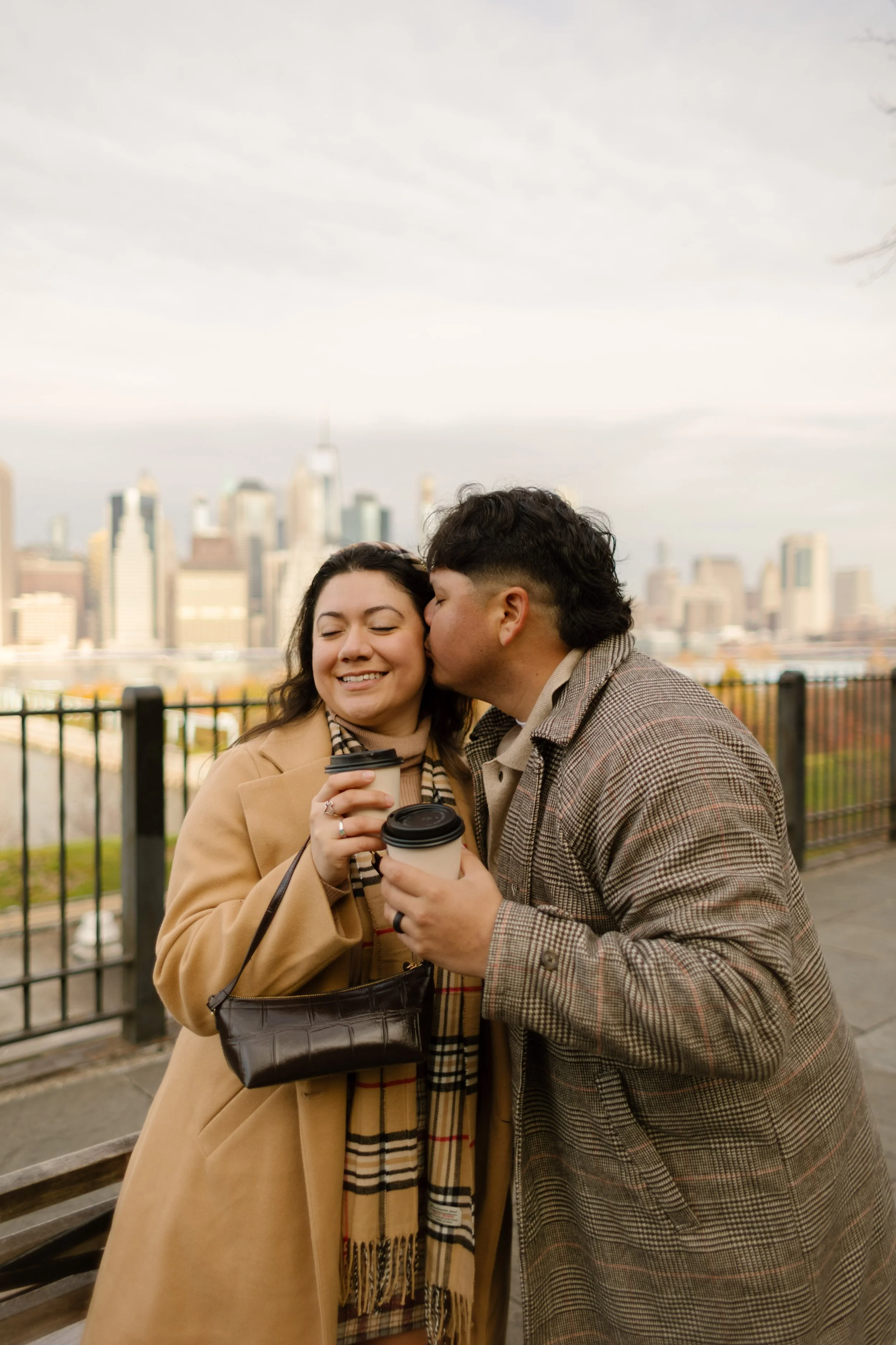 Candid New York engagement photo of a couple sharing coffee while one kisses the other on the cheek with the Manhattan skyline behind them.