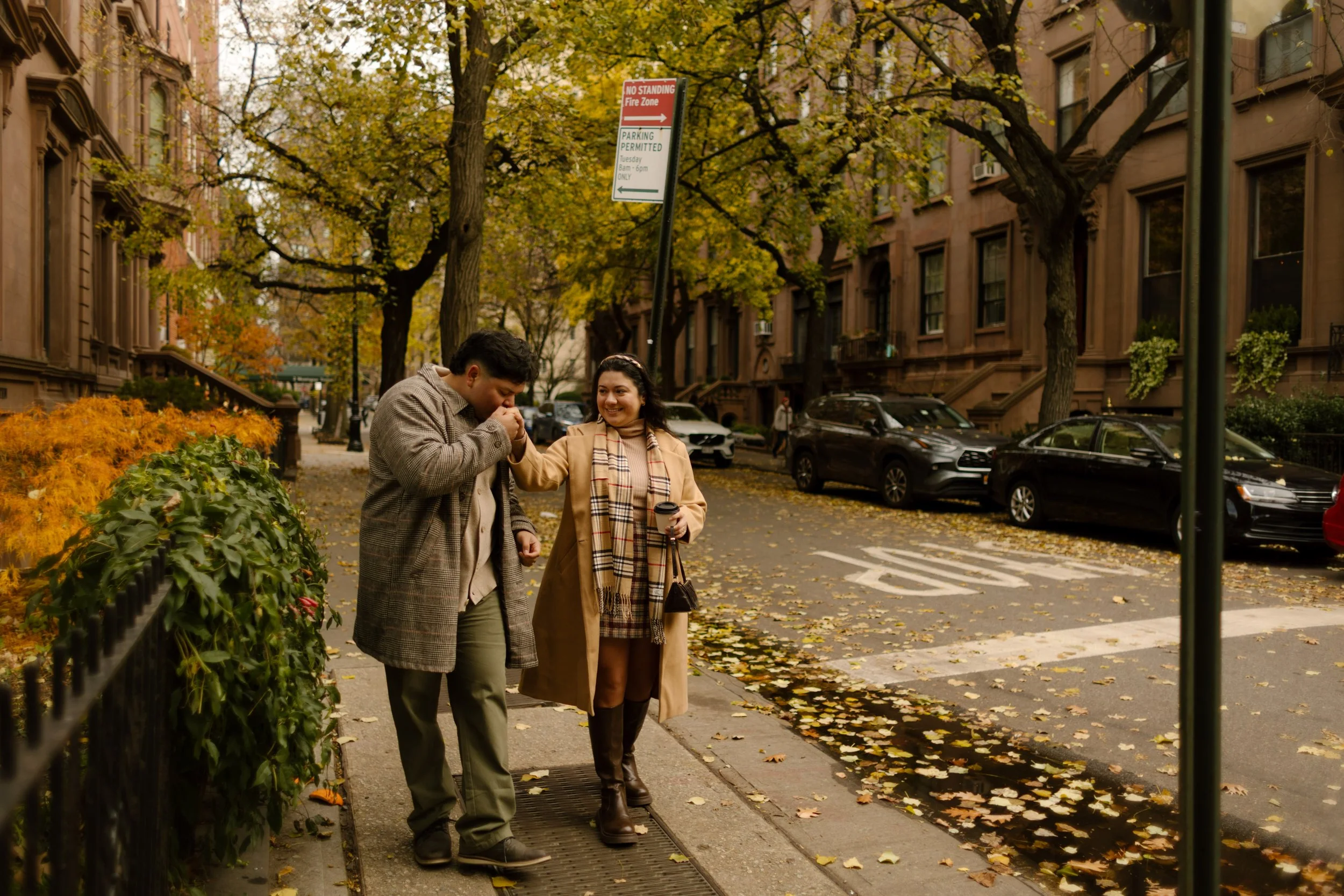 Brooklyn engagement photos of a couple walking through Brooklyn Heights as one kisses the other’s hand beneath colorful fall trees.