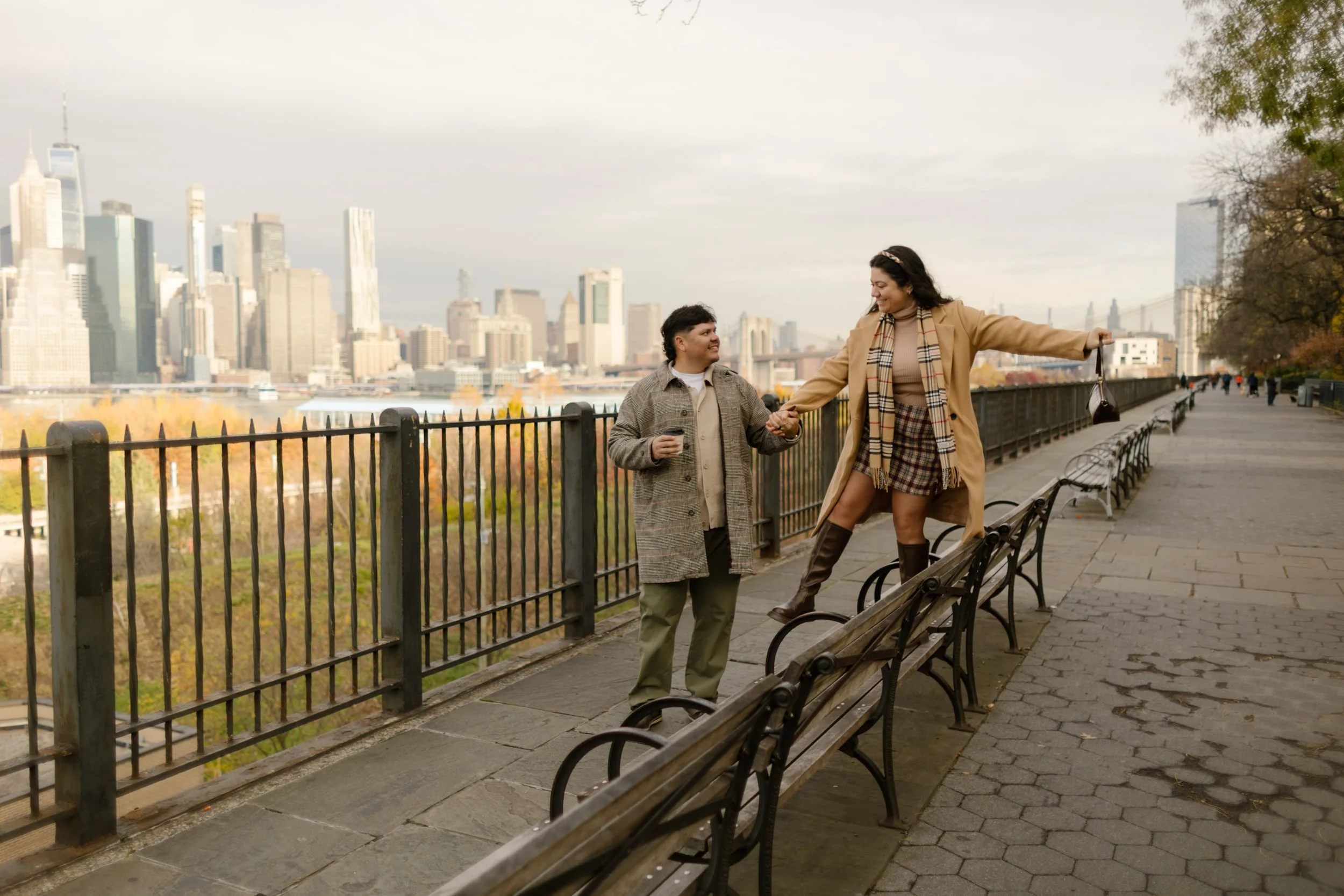 Playful fall engagement photo of a couple holding hands along the Brooklyn Heights Promenade with the Manhattan skyline behind them.