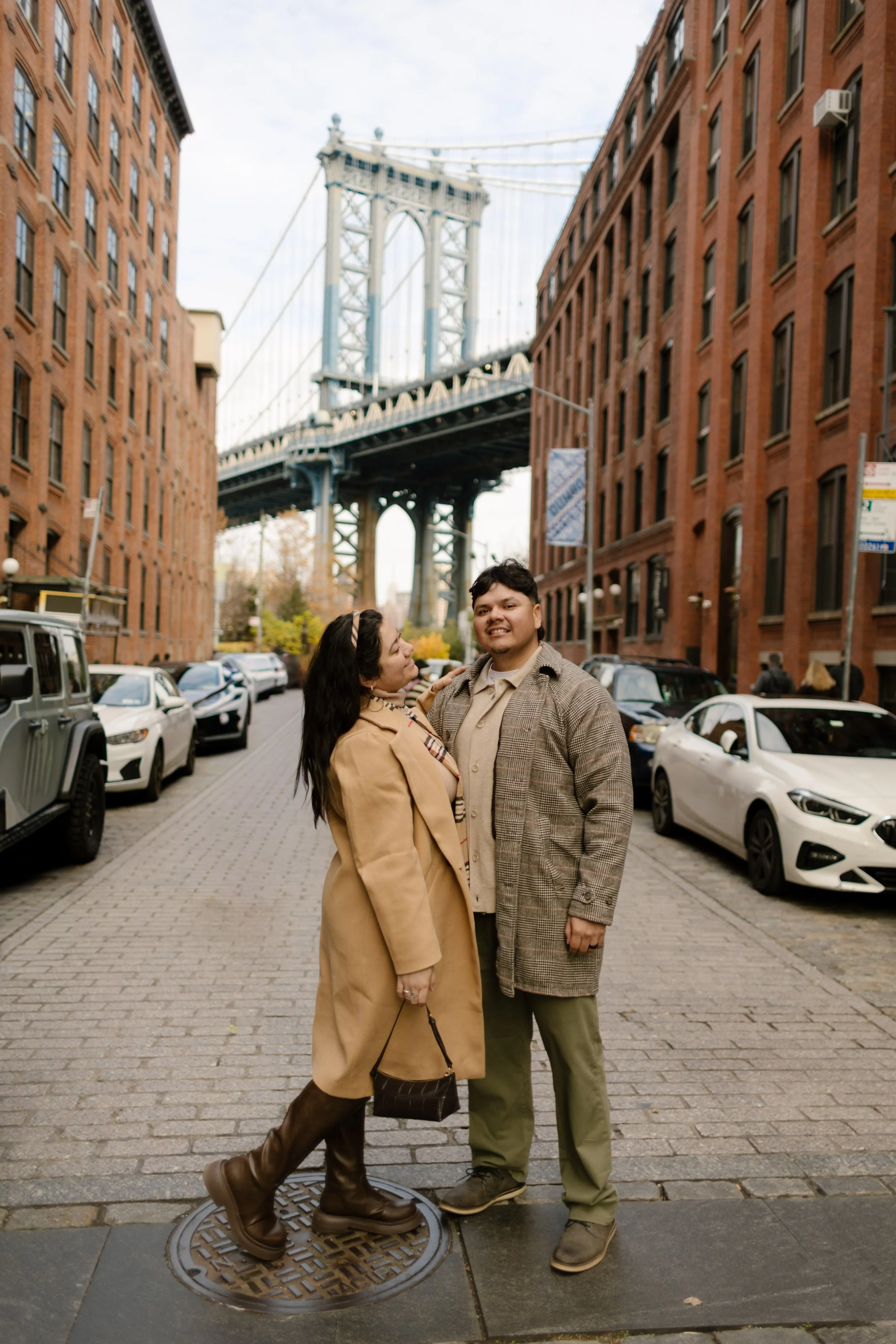 Brooklyn engagement photos of a couple posing on a cobblestone DUMBO street with the Manhattan Bridge and brick buildings behind them.