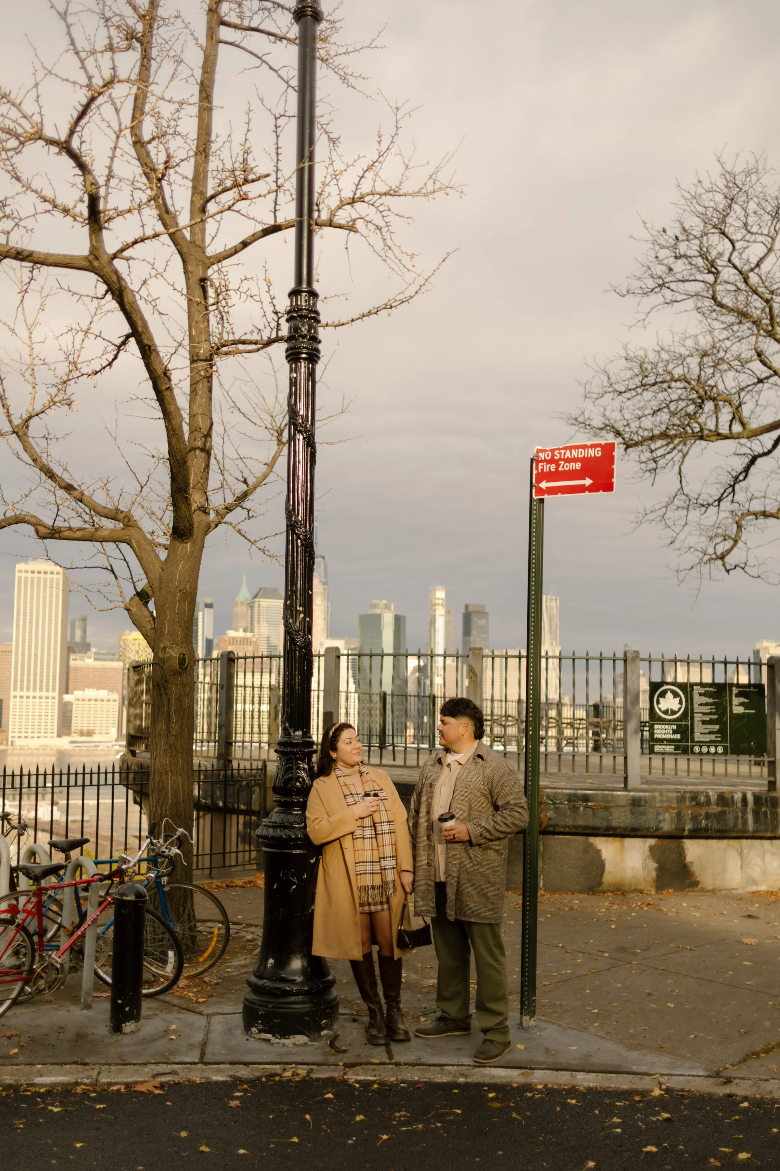 Brooklyn engagement photos of a couple standing together on a city corner with fall trees and the Manhattan skyline in the distance.