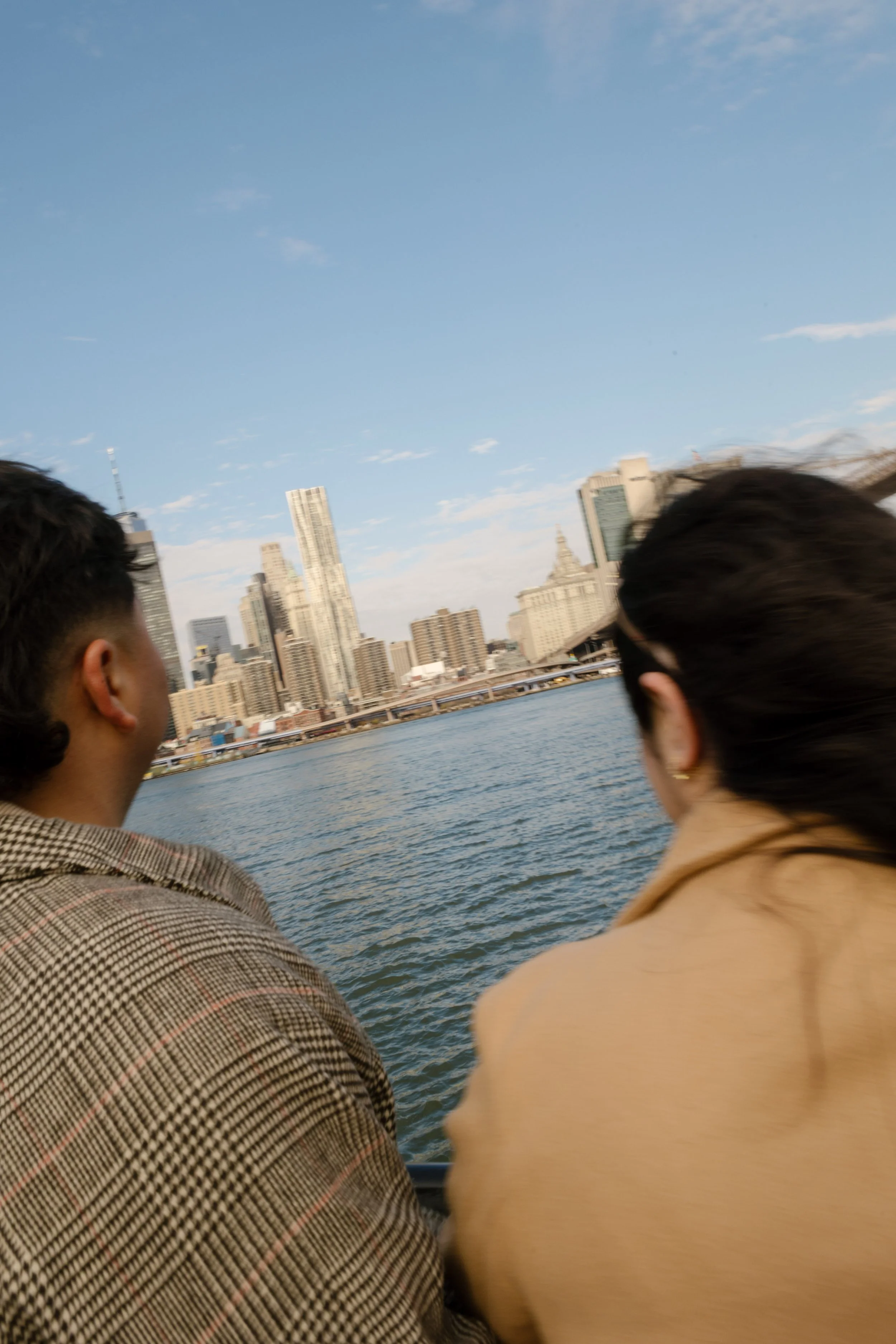 Candid waterfront engagement photo of a couple looking across the East River toward the Manhattan skyline.