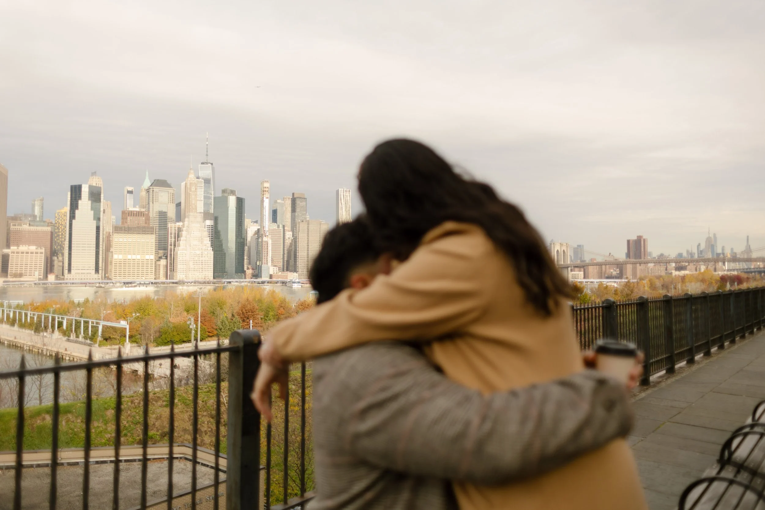 Brooklyn engagement photos of a couple hugging at a scenic overlook with the Manhattan skyline and fall foliage behind them.