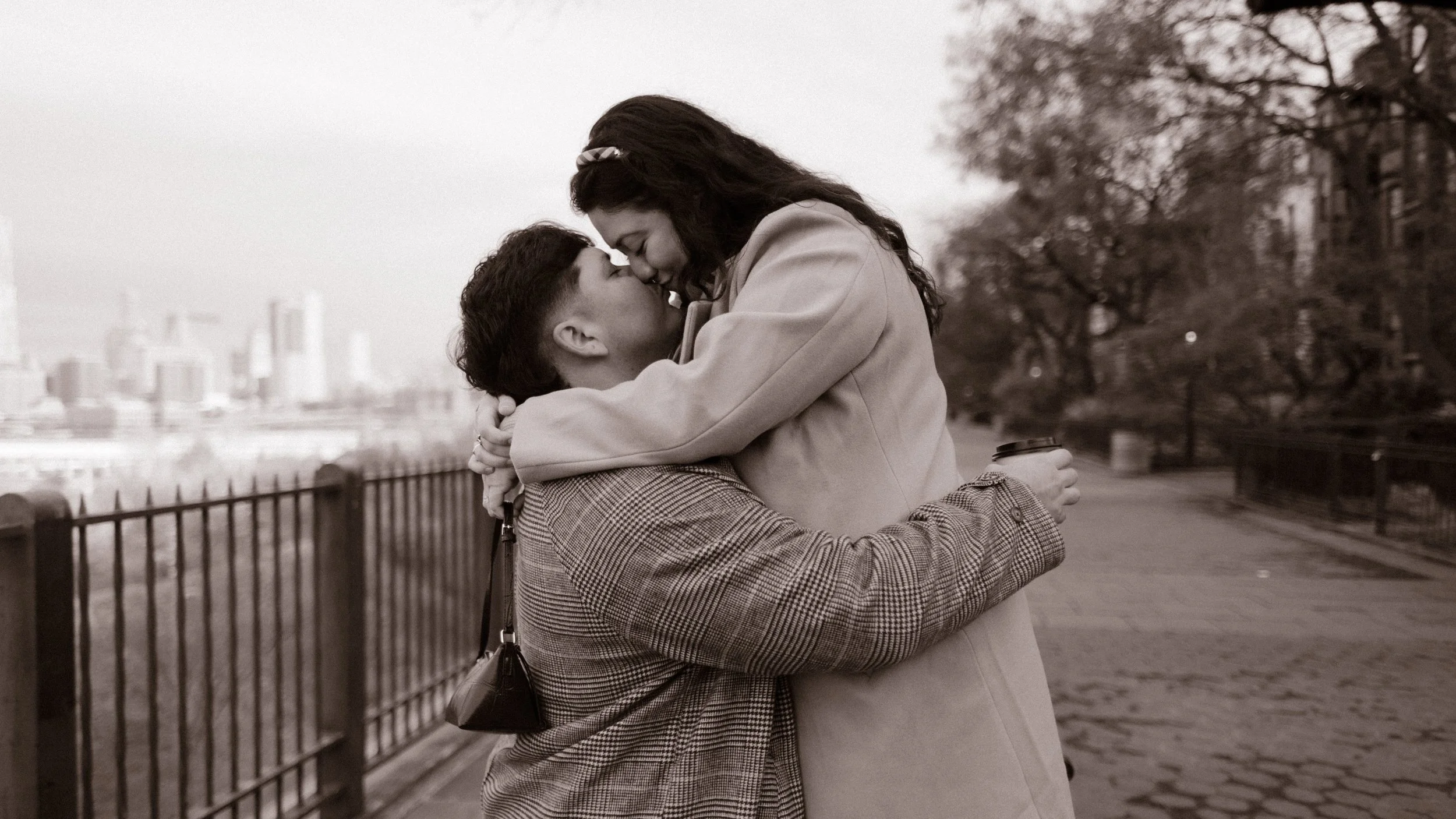 Black-and-white candid engagement photo of a couple laughing and embracing near the Brooklyn waterfront.